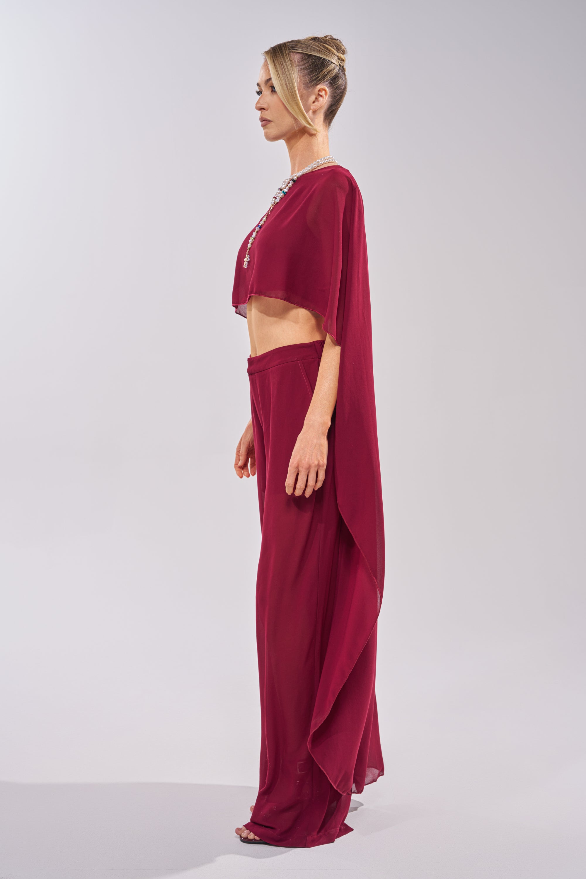 A woman in profile wears the COOL GIRL CHIFFON BLOUSE IN BURGUNDY, styled with wide-legged pants and a statement necklace. Her hair is in an elegant updo, set against a plain light background for easy day-to-night style. 3