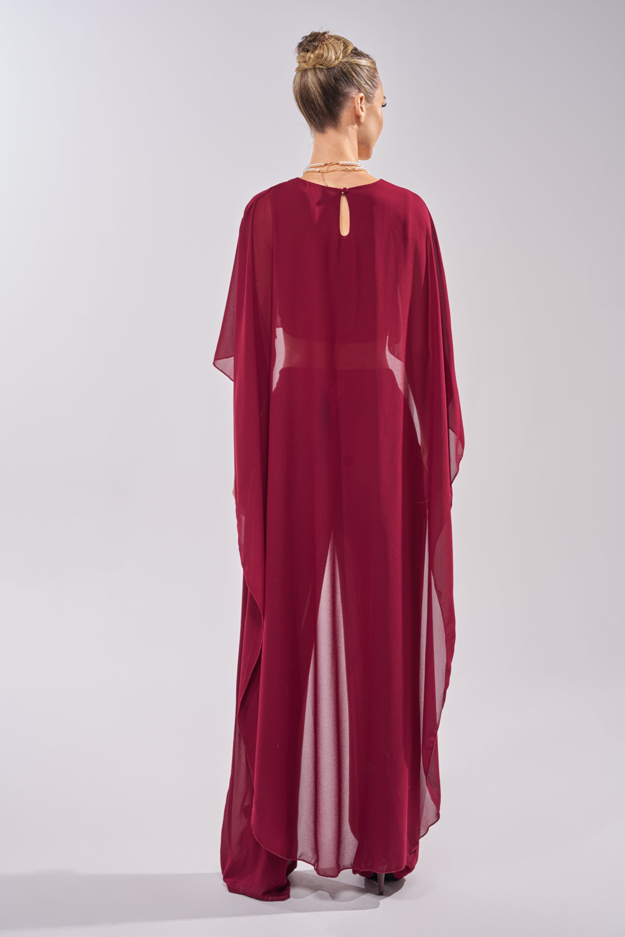 A woman with blonde hair in a bun models the COOL GIRL CHIFFON BLOUSE IN BURGUNDY, styled for day-to-night wear, standing with her back to the camera against a plain light gray background. 4