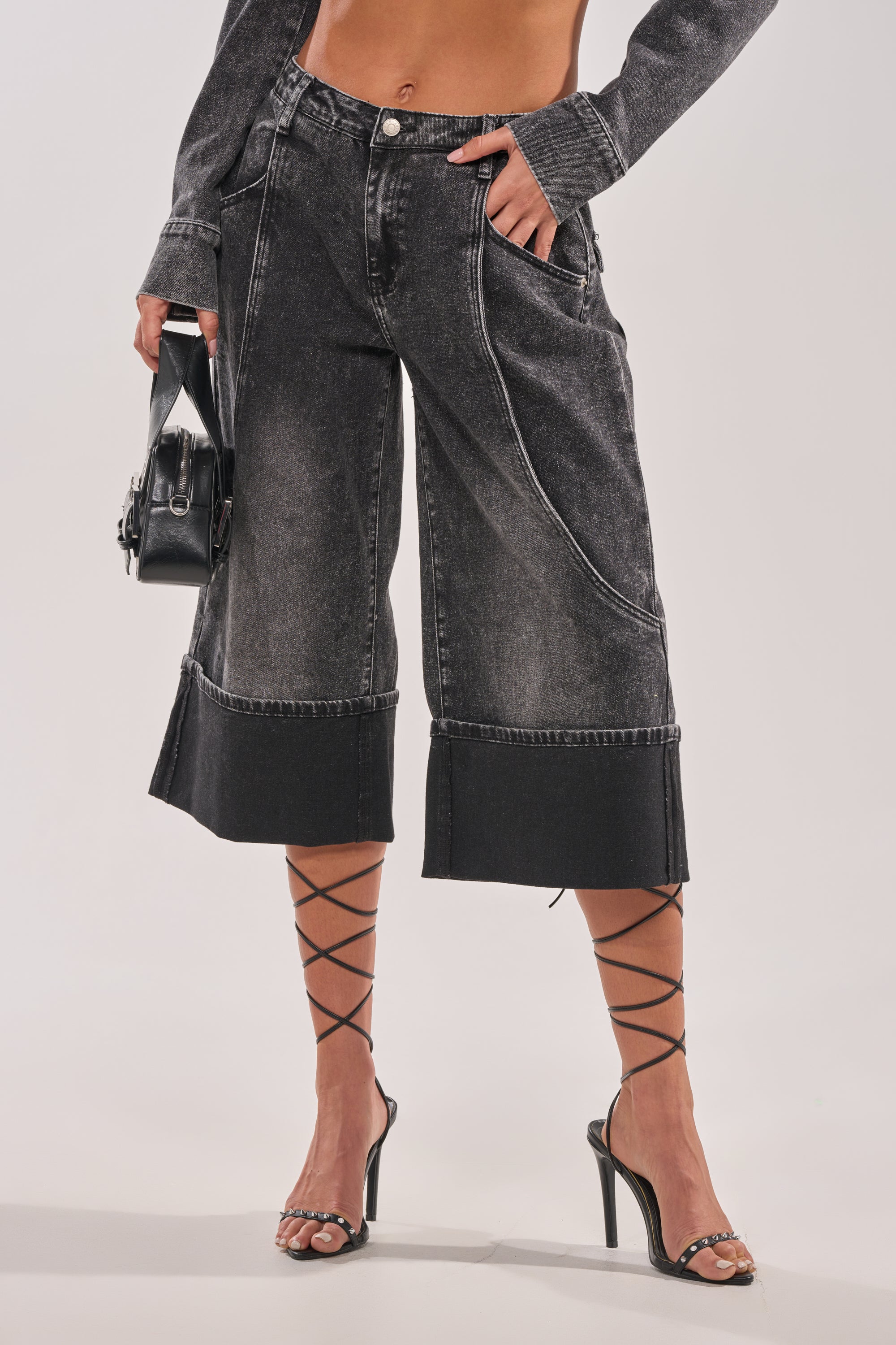 A person wearing the IN MY OWN WORLD BERMUDA SHORT with a matching denim jacket, black strappy high heels, and holding a small black handbag, shown from midriff to feet against a plain background. 1