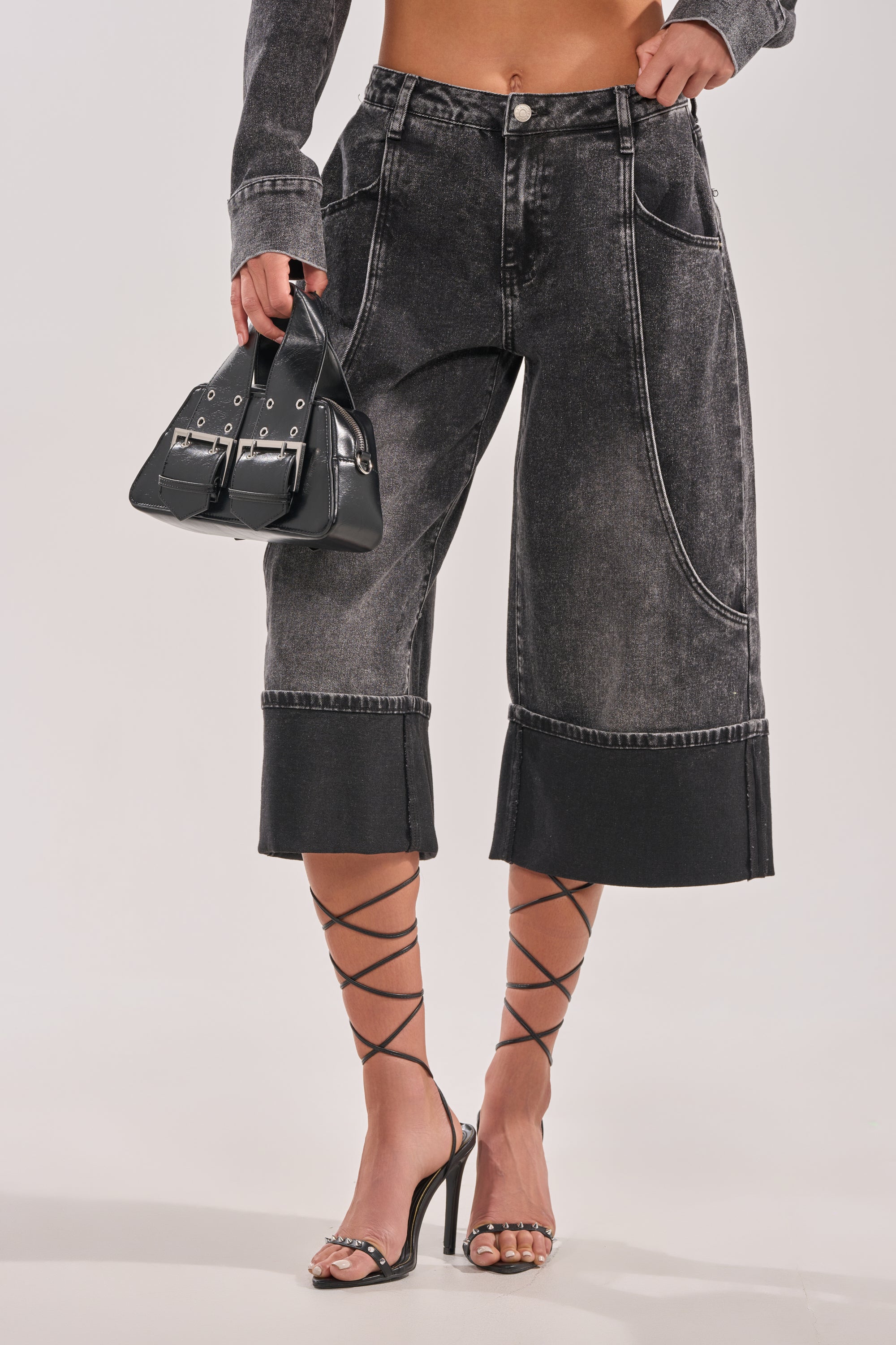 Wearing the IN MY OWN WORLD BERMUDA SHORT, a high-rise, baggy skater short, styled with a matching long-sleeve top, strappy high-heeled sandals, and a small black handbag against a plain background. 2