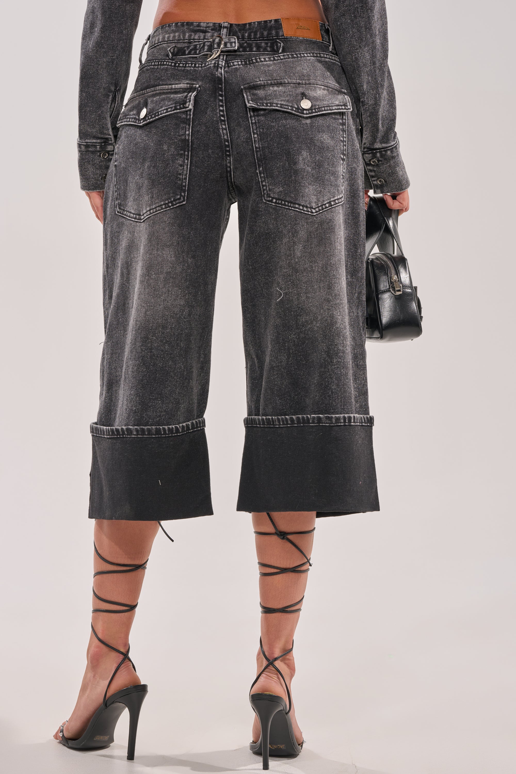 A person is seen from behind wearing the IN MY OWN WORLD BERMUDA SHORT, featuring a high-rise waist and visible seams, styled with strappy heeled sandals and carrying a small black purse. 4