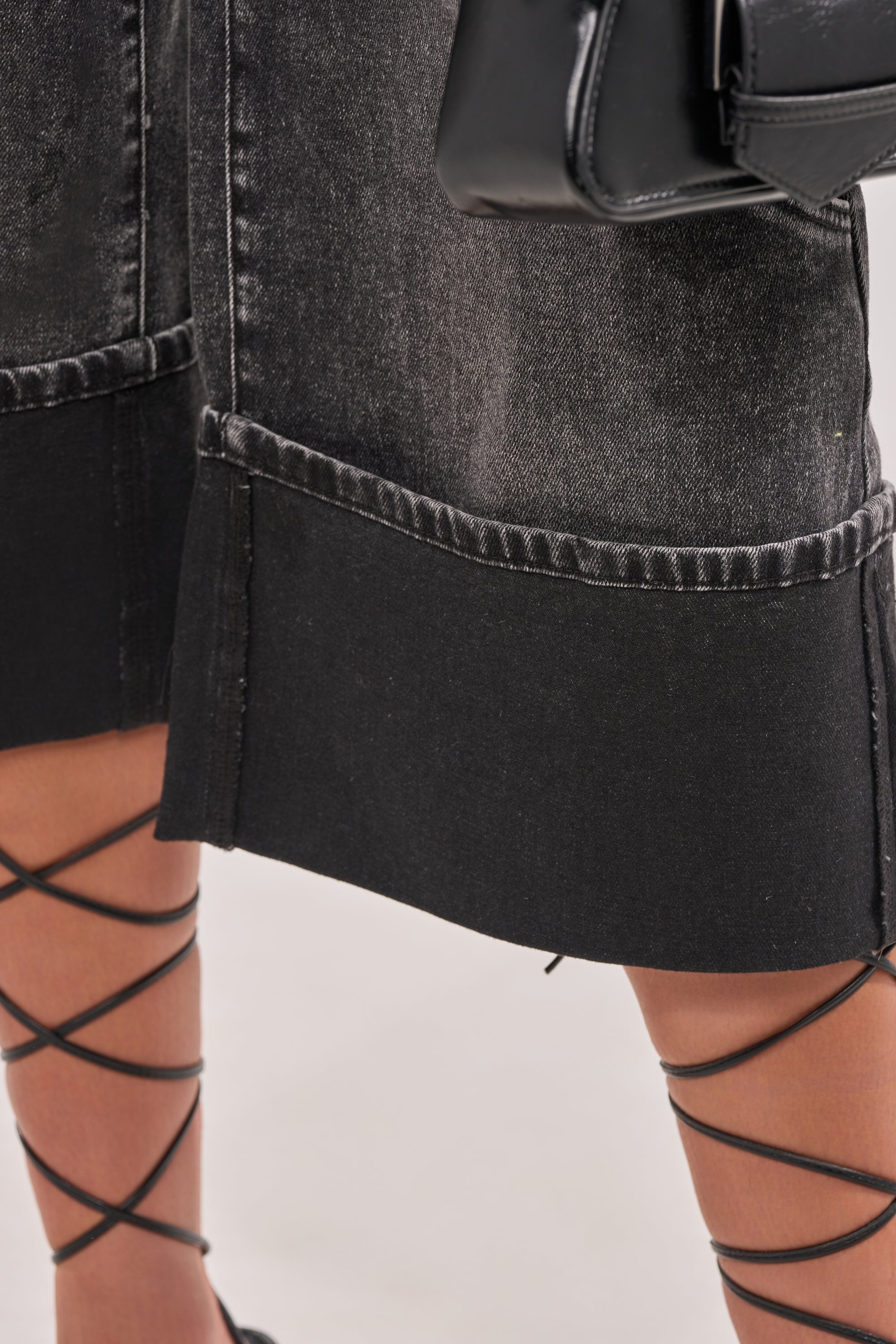 Close-up of a person wearing the IN MY OWN WORLD BERMUDA SHORT—black denim bermuda shorts with wide, cuffed hems—styled with black lace-up sandals wrapped around the ankles and lower legs. 7