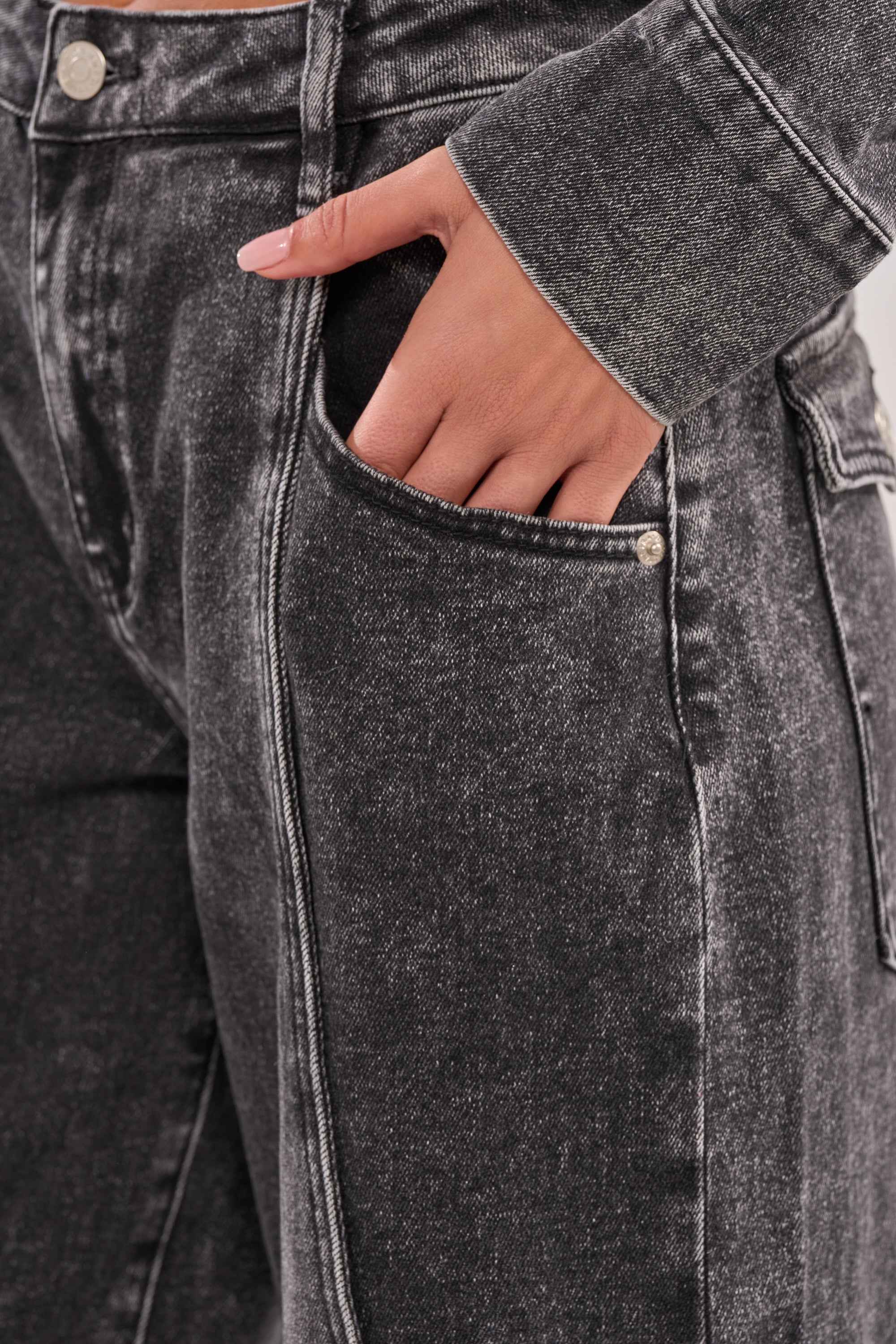 A close-up of a person wearing the IN MY OWN WORLD BERMUDA SHORT—a black, high-rise denim bermuda—with a matching jacket. Their light pink manicured nails rest partially in the front pocket. 8