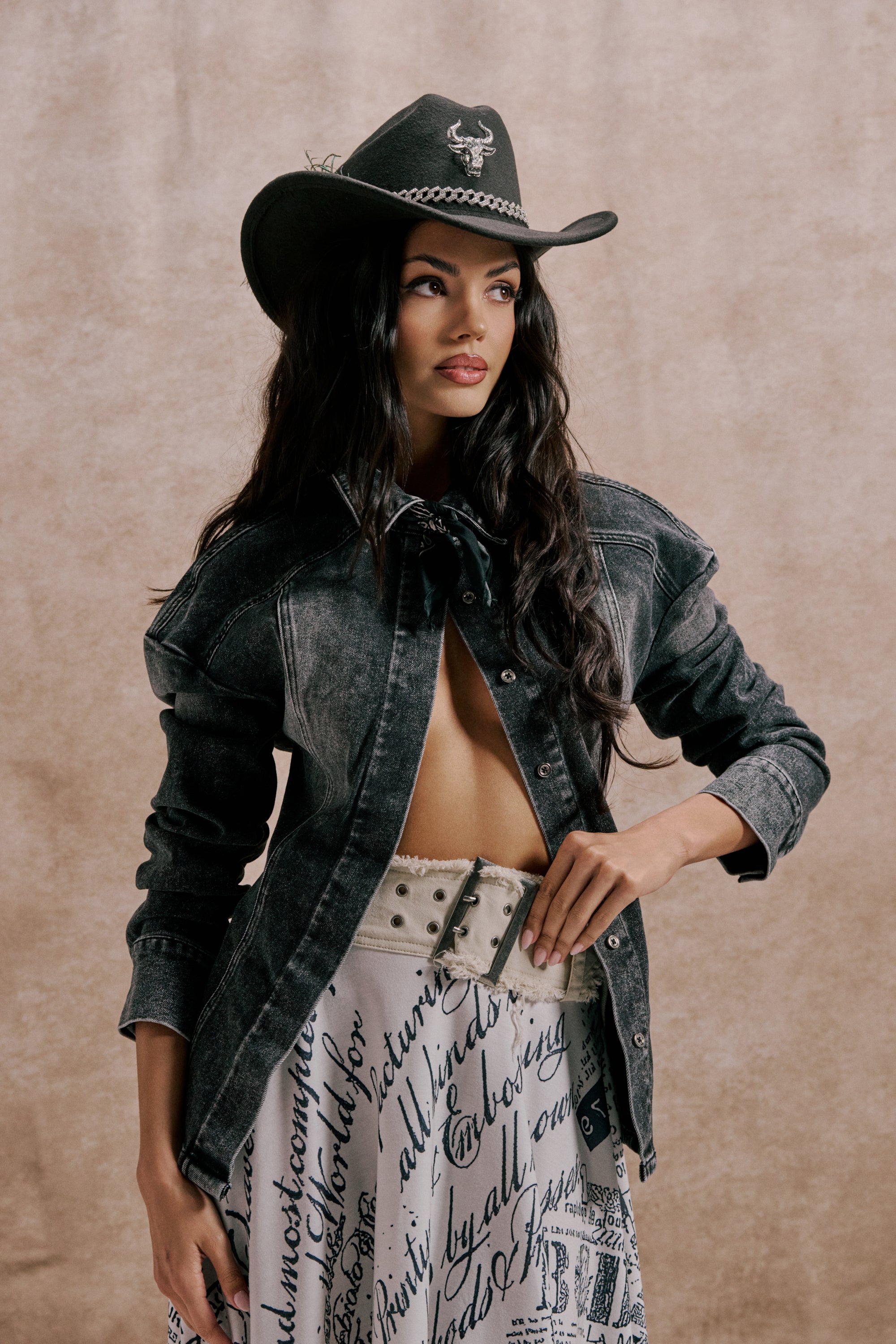 A woman in a black cowboy hat and washed black denim IN MY OWN WORLD BUTTON DOWN poses confidently in a white skirt with bold black script and a wide belt, standing against a neutral textured background. 2