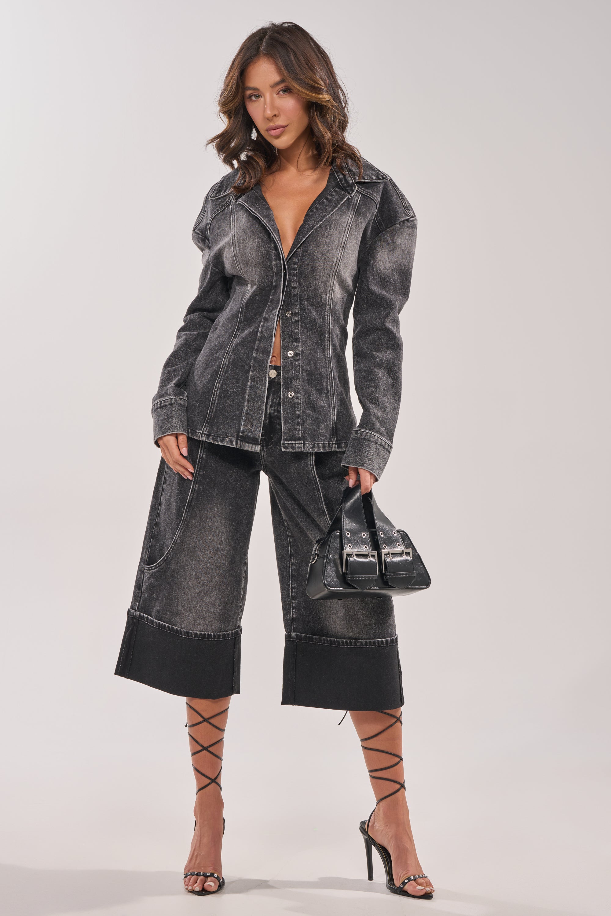 A woman wears the IN MY OWN WORLD BUTTON DOWN in washed black denim and matching wide-leg culottes, styled with a black adjustable-strap handbag and strappy high heels, standing against a light background with wavy hair. 9