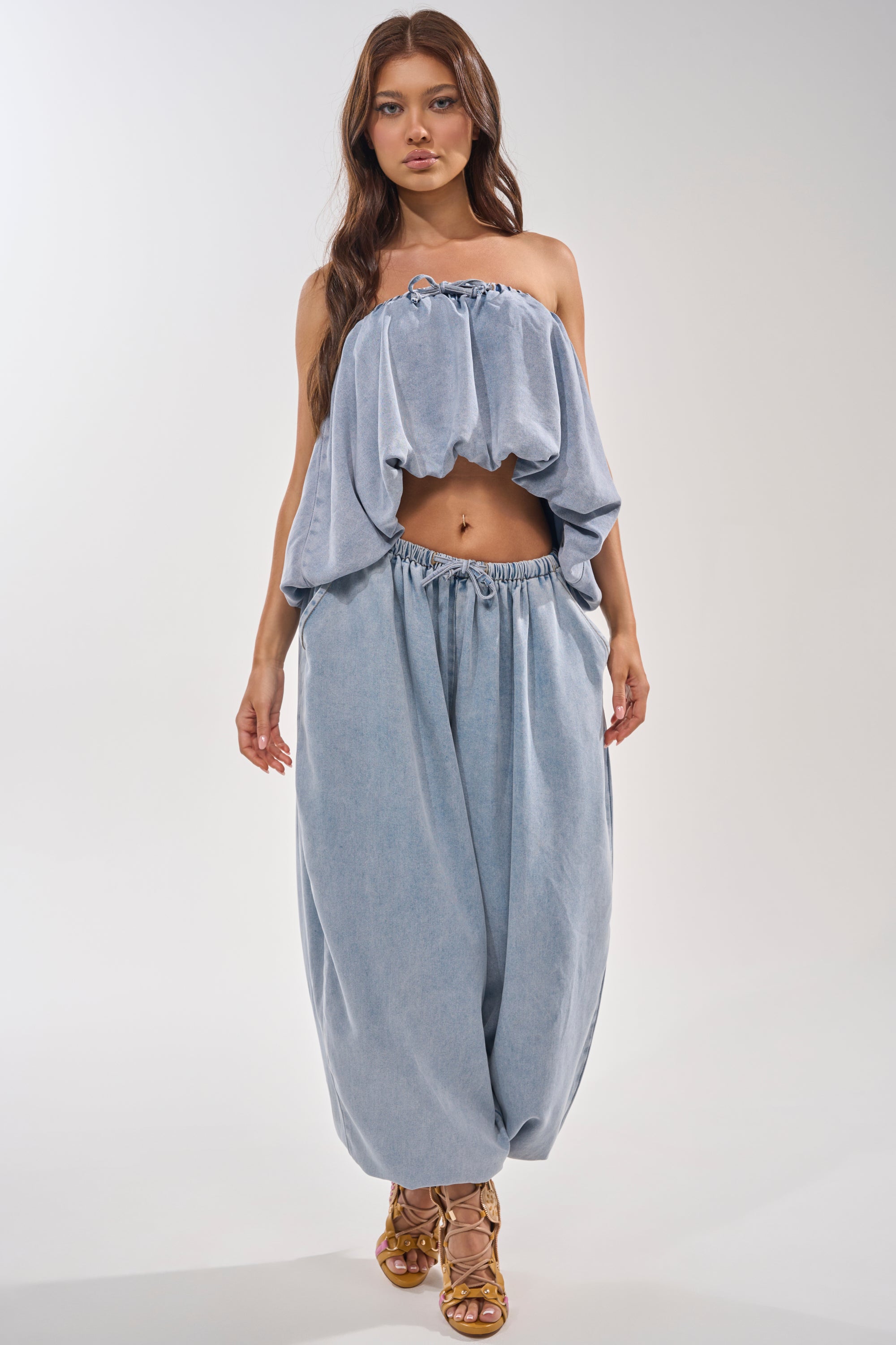 A woman with long brown hair wears the PUMPED UP DENIM BLOUSE, a light wash tube top, paired with matching wide, baggy pants and tan strappy sandals, standing against a plain white background. 6