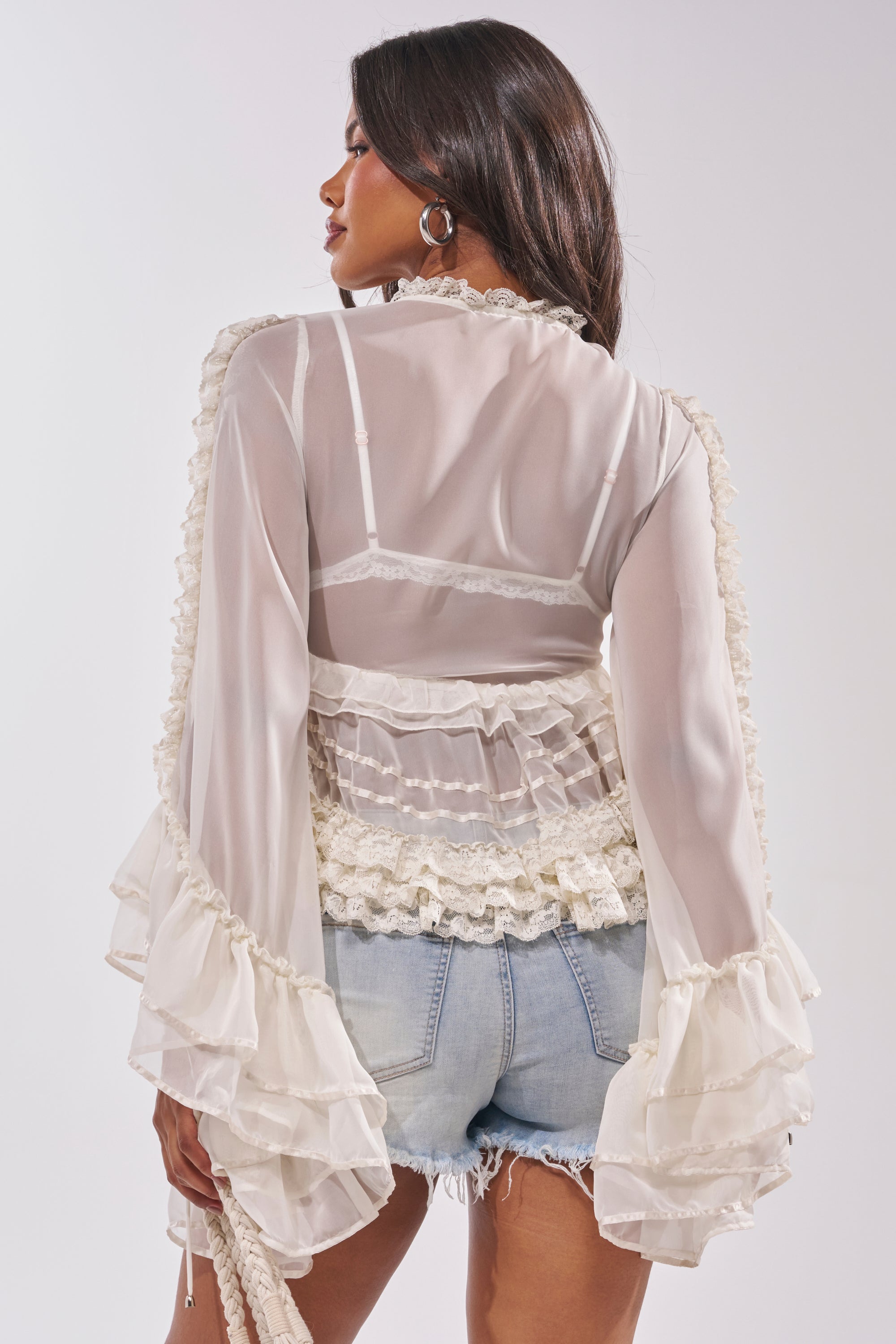 A woman with long dark hair models the BRIDGETTE LONG SLEEVE BLOUSE, featuring sheer white ruffles and flared sleeves, paired with light denim cutoff shorts. She is turned away to highlight the blouse's back details against a plain light background. 2