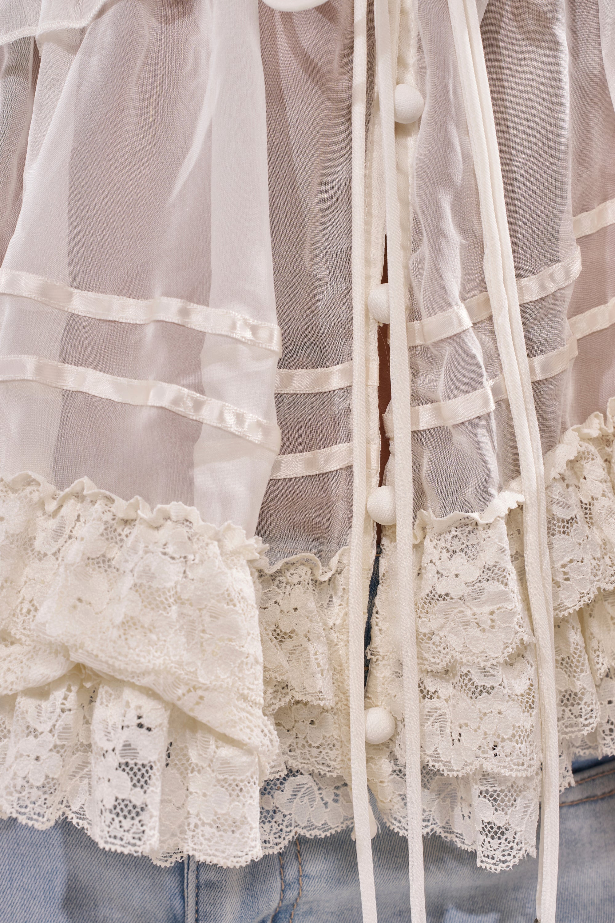 Close-up of the BRIDGETTE LONG SLEEVE BLOUSE, featuring sheer white fabric, lace trim, and fabric-covered buttons, styled effortlessly with light blue denim jeans for a chic look. 6