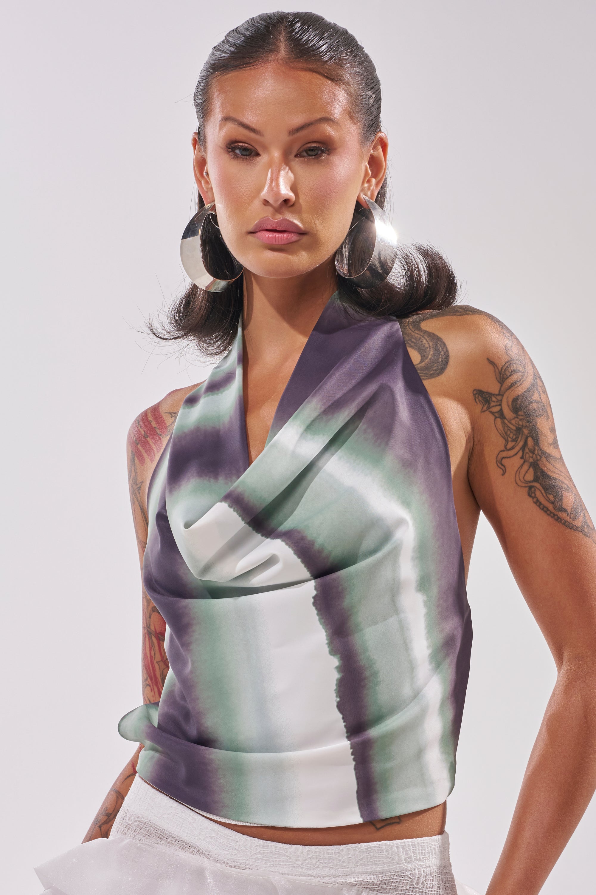 A woman with large hoop earrings and visible tattoos models the TALIA OPEN BACK COWL NECK TOP, a sleeveless satin piece in a green-and-navy print, standing against a simple light backdrop. 0