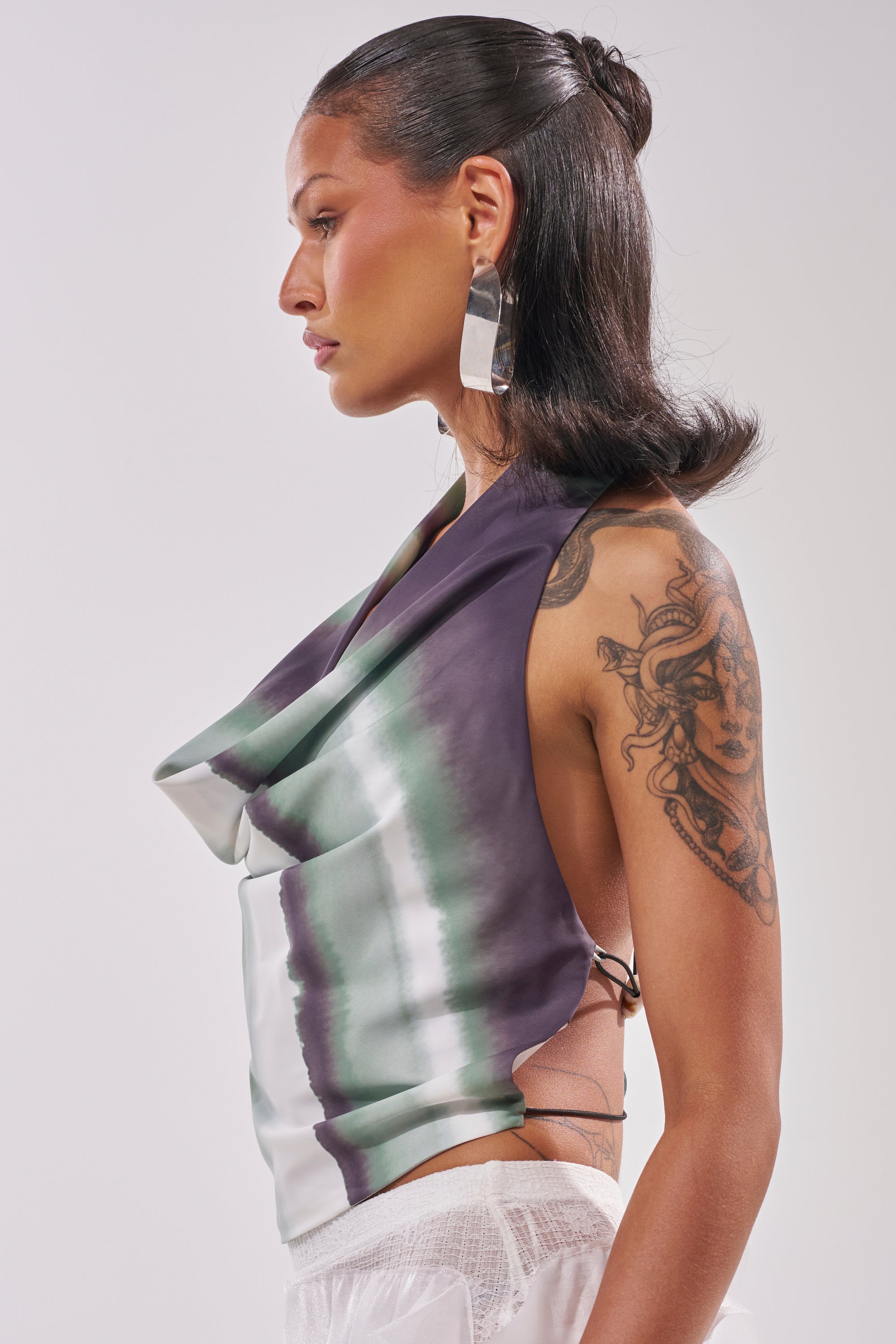 A woman in profile models the TALIA OPEN BACK COWL NECK TOP—a summer-chic, sleeveless satin blouse with green, black, and white tie-dye. She wears long earrings, has sleek dark hair, and displays a large tattoo of a woman's face on her upper arm. 2