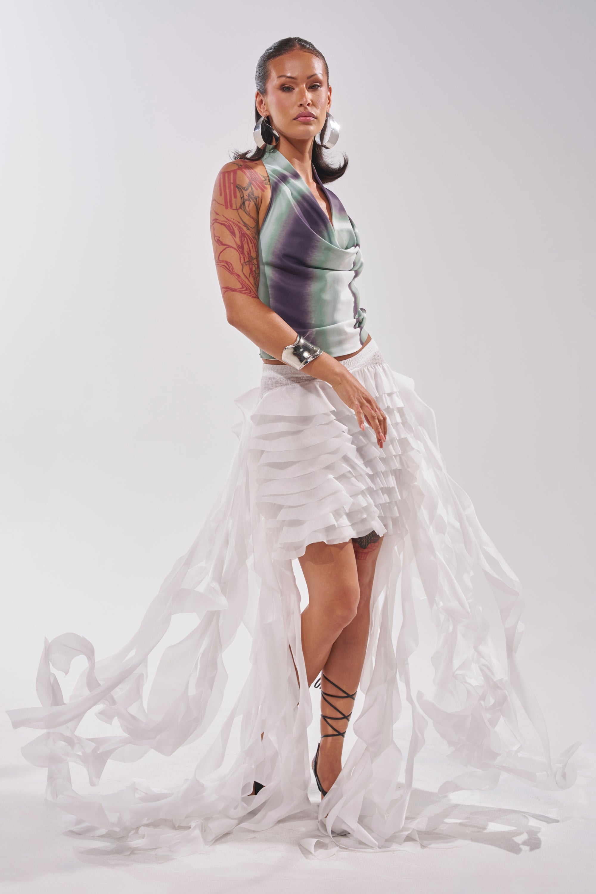 A woman with dark hair, tattoos, hoop earrings, a silver cuff, and strappy black heels stands confidently in the TALIA OPEN BACK COWL NECK TOP and a flowing white layered skirt with ribbon-like strips. 4