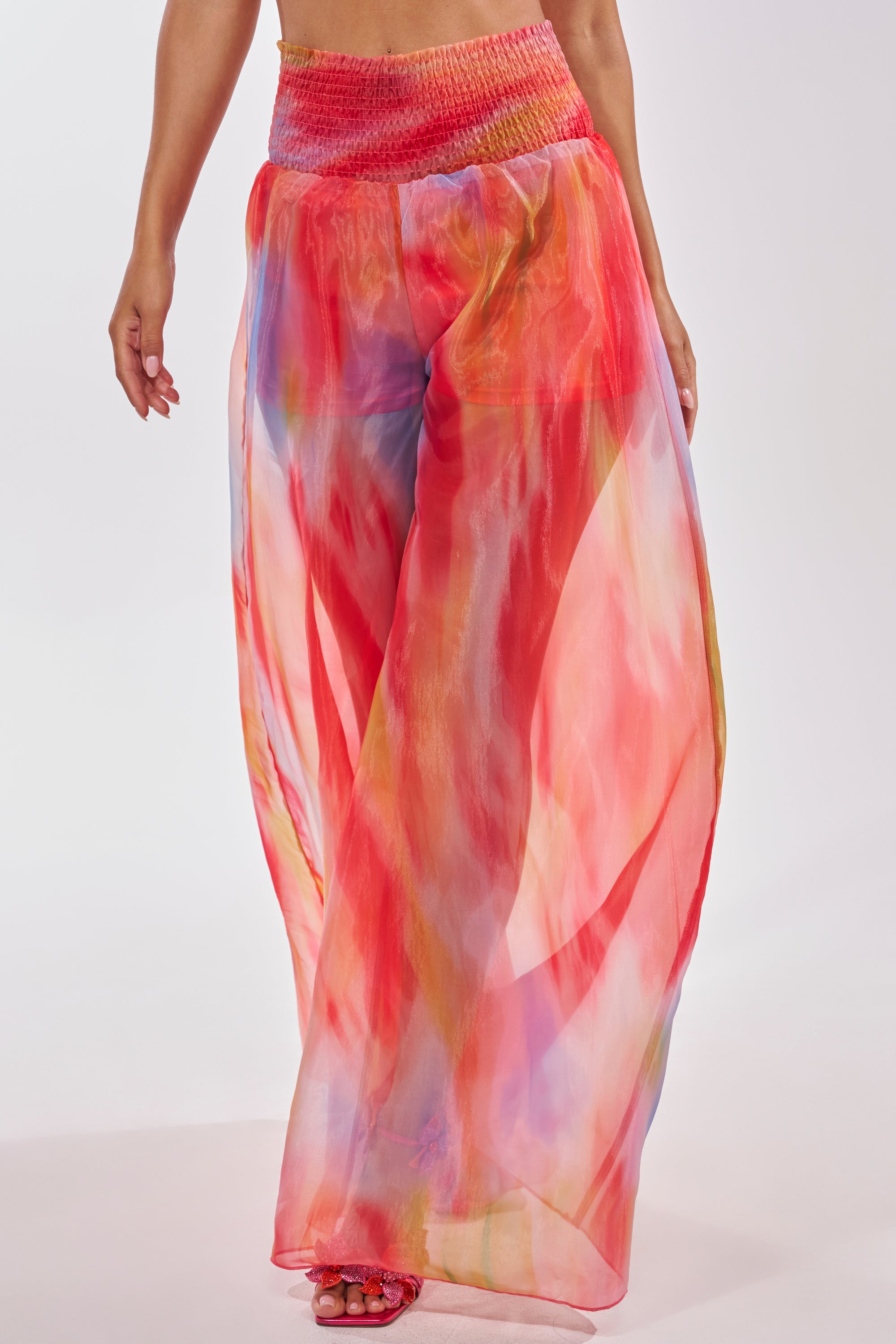 A person wears the INTERESTED IN YOU PALAZZO PANT, featuring sheer organza with a watercolor print in pink, red, orange, and blue, a red smocked waistband, wide-leg silhouette, and open-toe sandals. 0