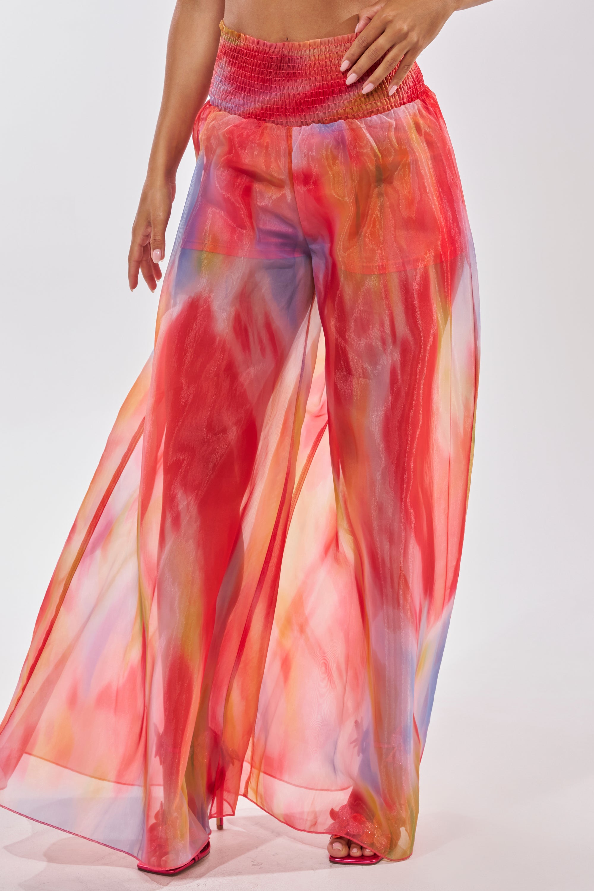 A person wears the INTERESTED IN YOU PALAZZO PANT—high-rise, wide-leg sheer organza pants with a smocked waistband and a vibrant red, pink, yellow, and blue watercolor print. Only the lower body is visible against a plain background. 1
