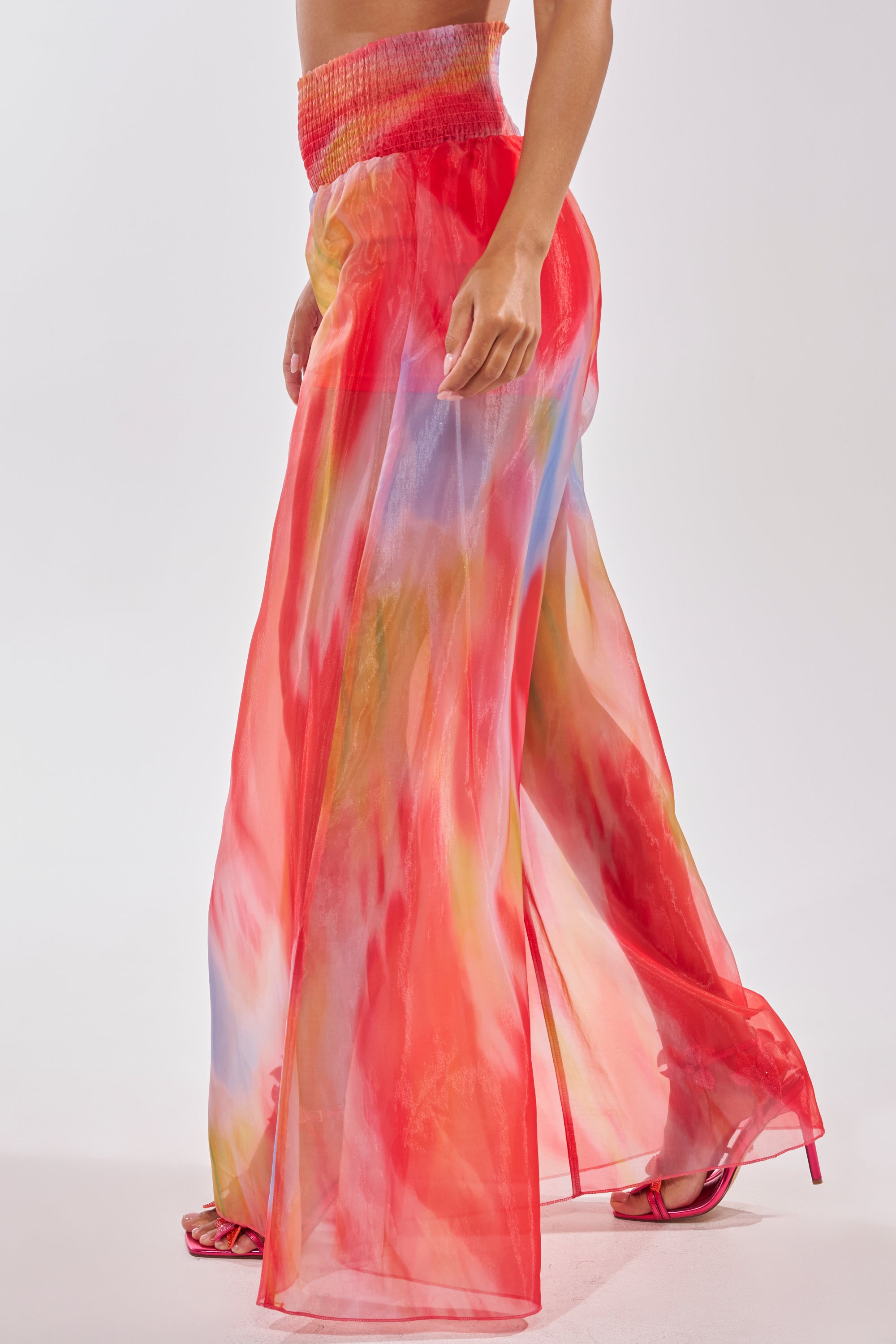 A person wears the INTERESTED IN YOU PALAZZO PANT, a high-waisted organza pant with a sheer red, yellow, and blue watercolor print, paired with red high-heeled sandals against a plain white background. 2