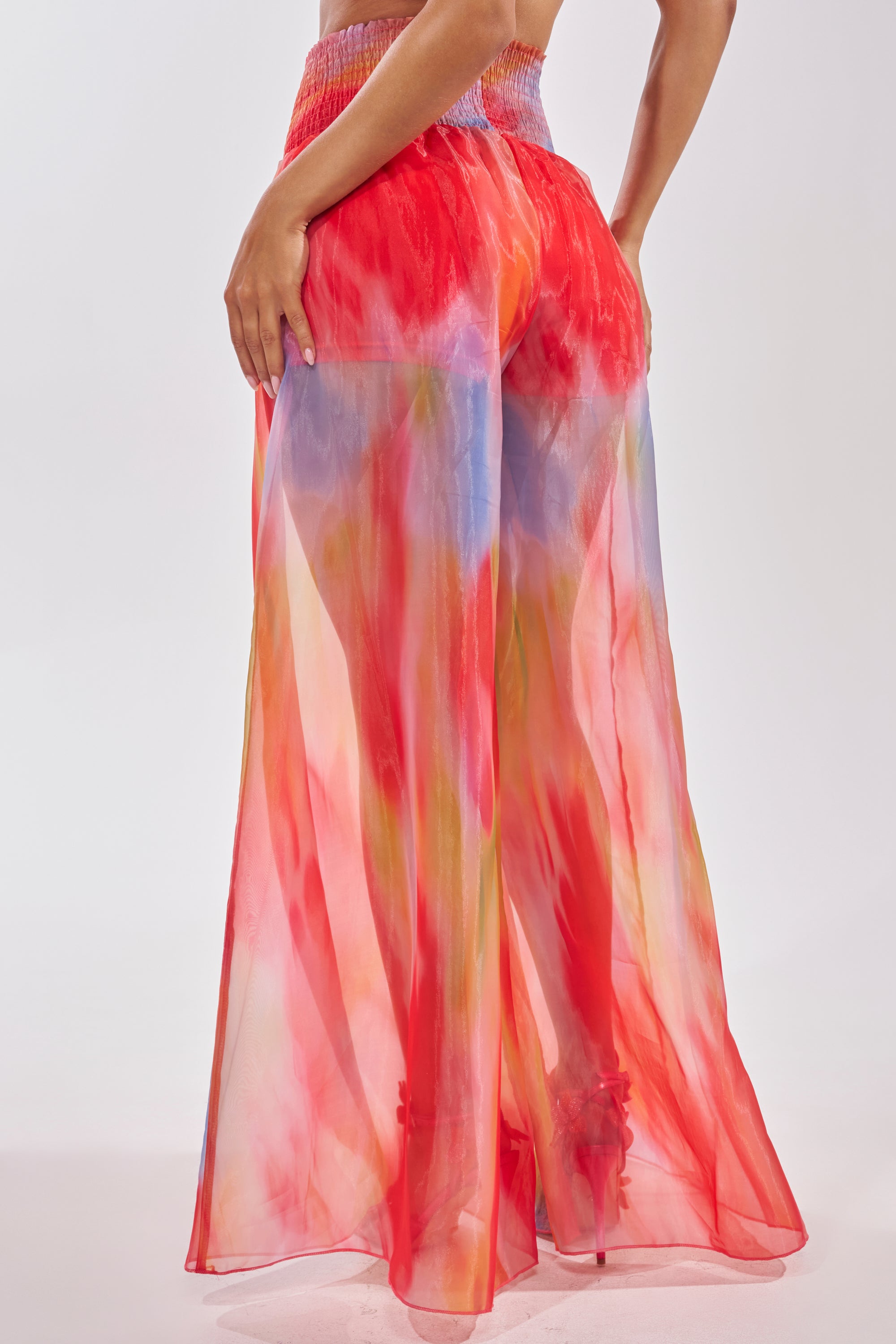Wearing the "INTERESTED IN YOU PALAZZO PANT," a person stands against a plain light background in high-rise, wide-leg organza pants featuring a vibrant red, pink, yellow, and blue watercolor print. 3
