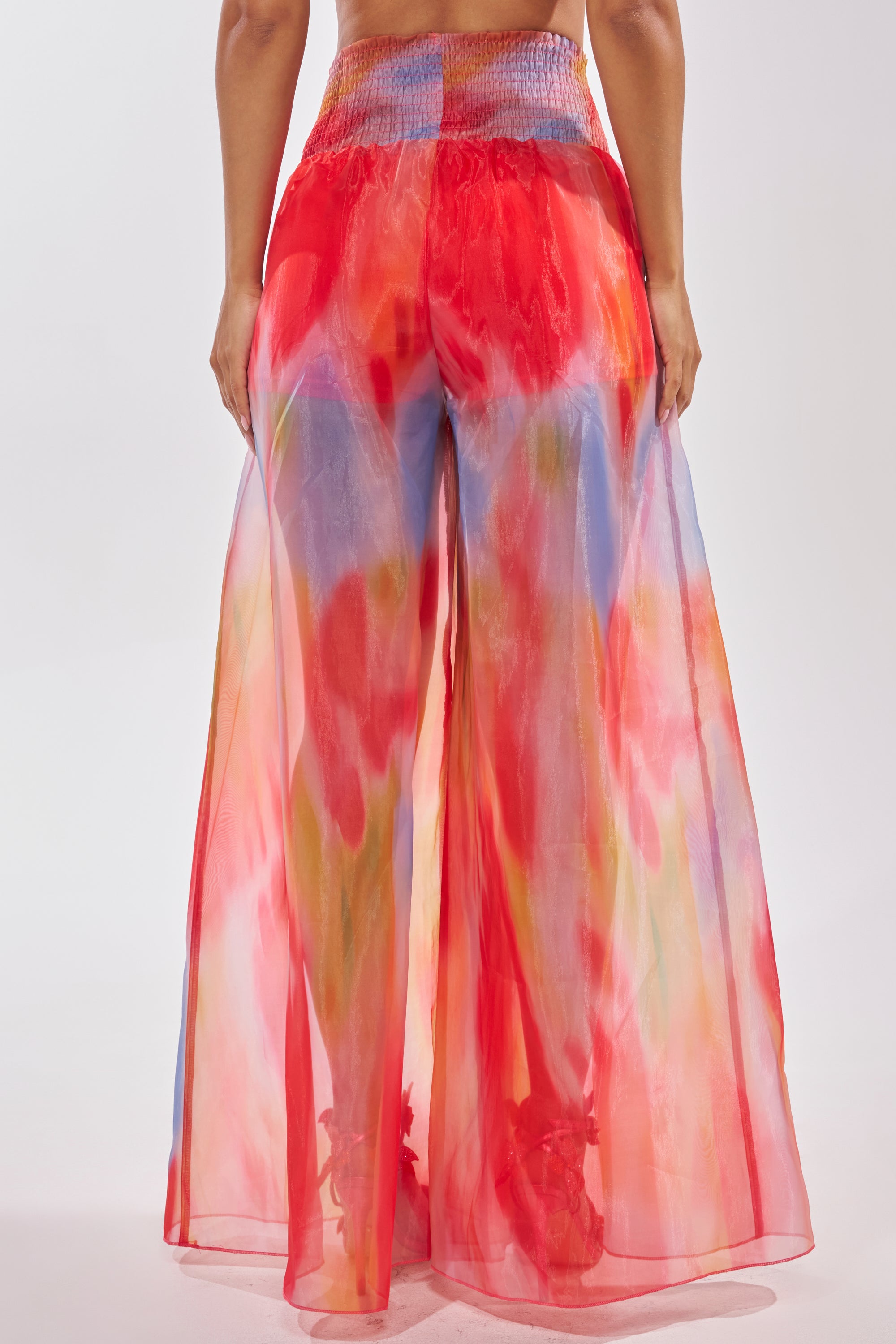 A person wearing the INTERESTED IN YOU PALAZZO PANT—high-rise, wide-leg organza pants with a colorful watercolor print in red, orange, blue, and yellow—viewed from the back against a plain light background. 4