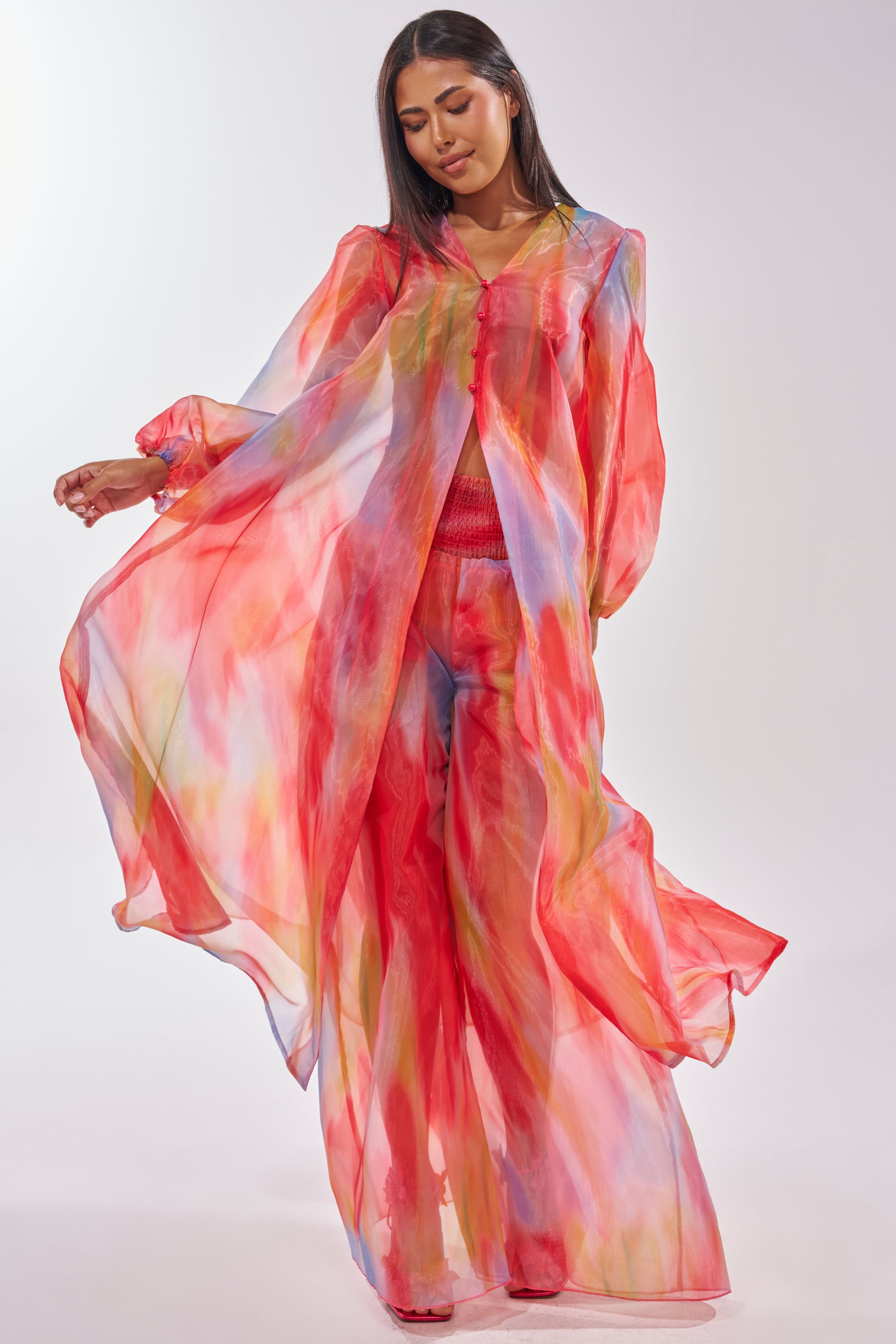 A woman poses gracefully with arms out and eyes closed, wearing the INTERESTED IN YOU DUSTER—a flowing, sheer organza piece featuring a watercolor wash of red, blue, and yellow—against a plain light background. 0