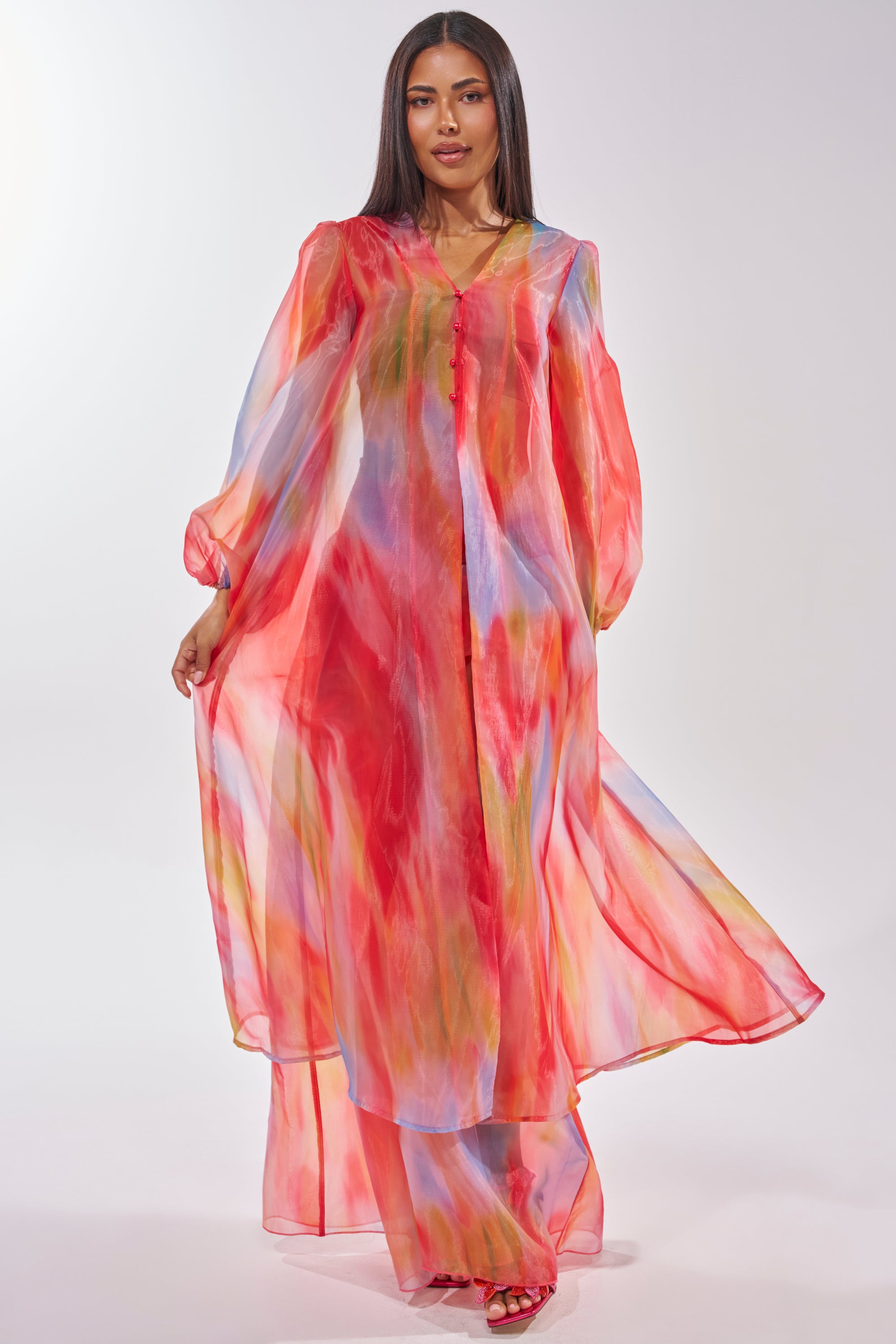 A woman with straight hair wears the INTERESTED IN YOU DUSTER, a sheer, flowing longline maxi dress featuring a colorful watercolor wash in red, blue, yellow, and pink. She completes the look with red sandals. 1
