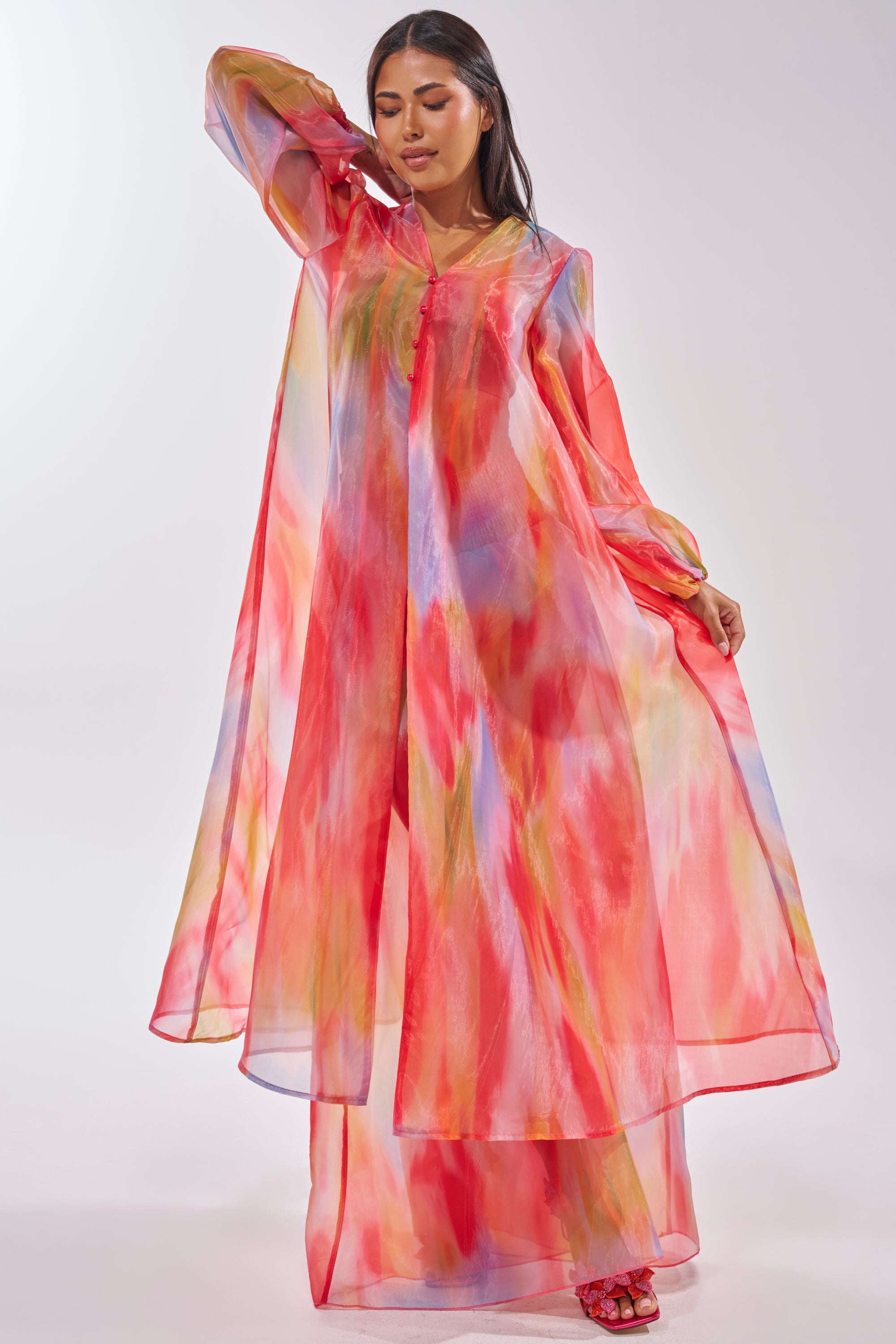 A woman wears the INTERESTED IN YOU DUSTER, a longline, sheer organza duster featuring multicolored shades of red, pink, yellow, and blue. She stands with one hand raised to her head and the other holding out the flowing fabric. 2