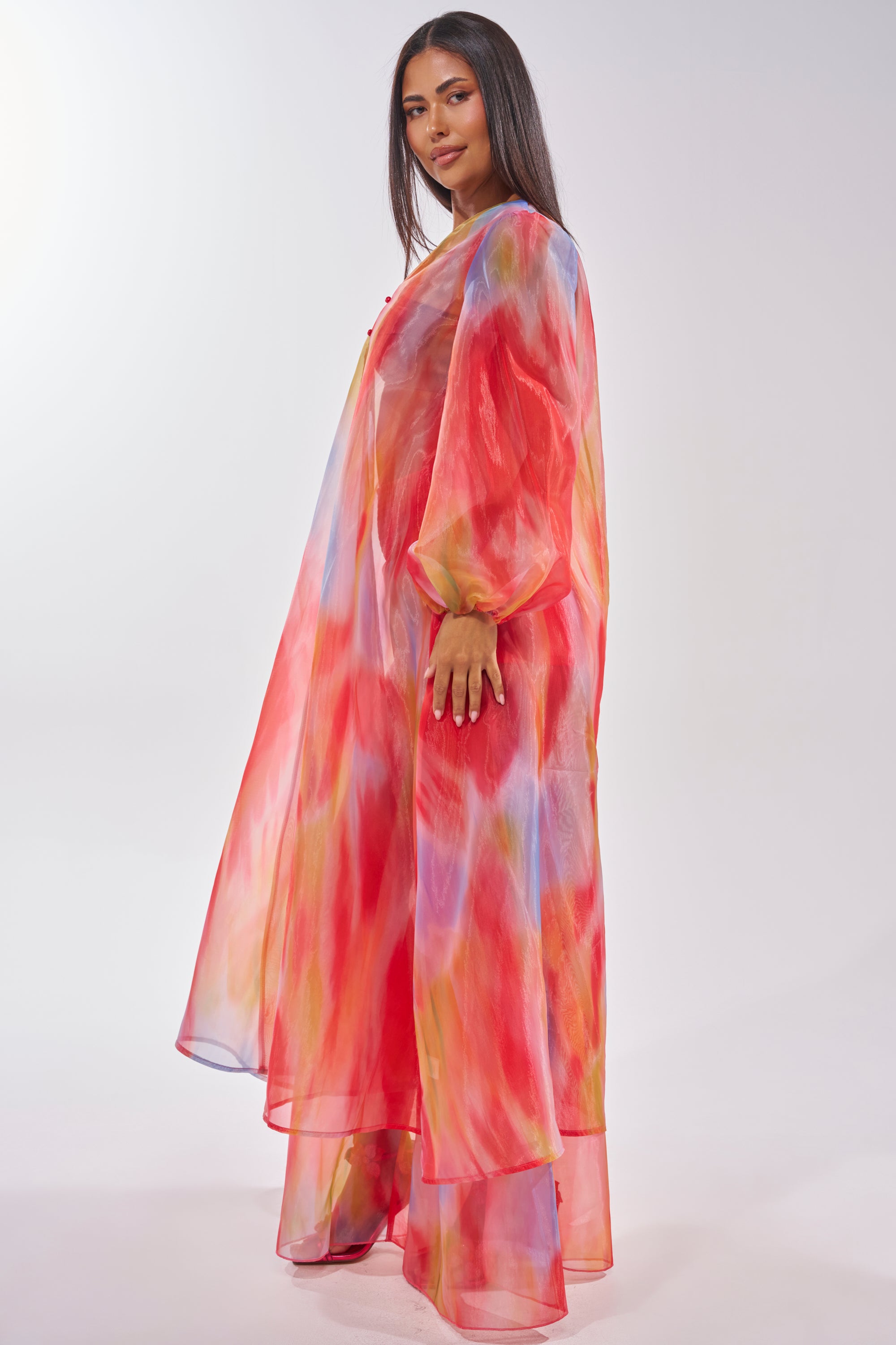 A woman stands in profile wearing the INTERESTED IN YOU DUSTER, a sheer organza piece with a longline silhouette and a vibrant watercolor pattern in red, pink, blue, and yellow. The backdrop is plain and well-lit. 4