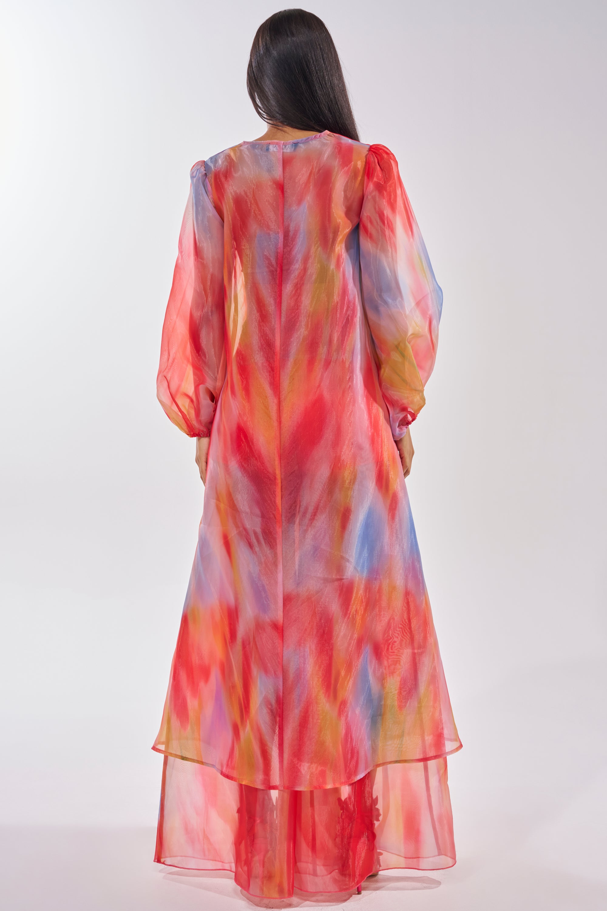 A woman with long dark hair is shown from the back, wearing the INTERESTED IN YOU DUSTER—a sheer organza duster with long sleeves, featuring a colorful abstract watercolor pattern on a white background. 5