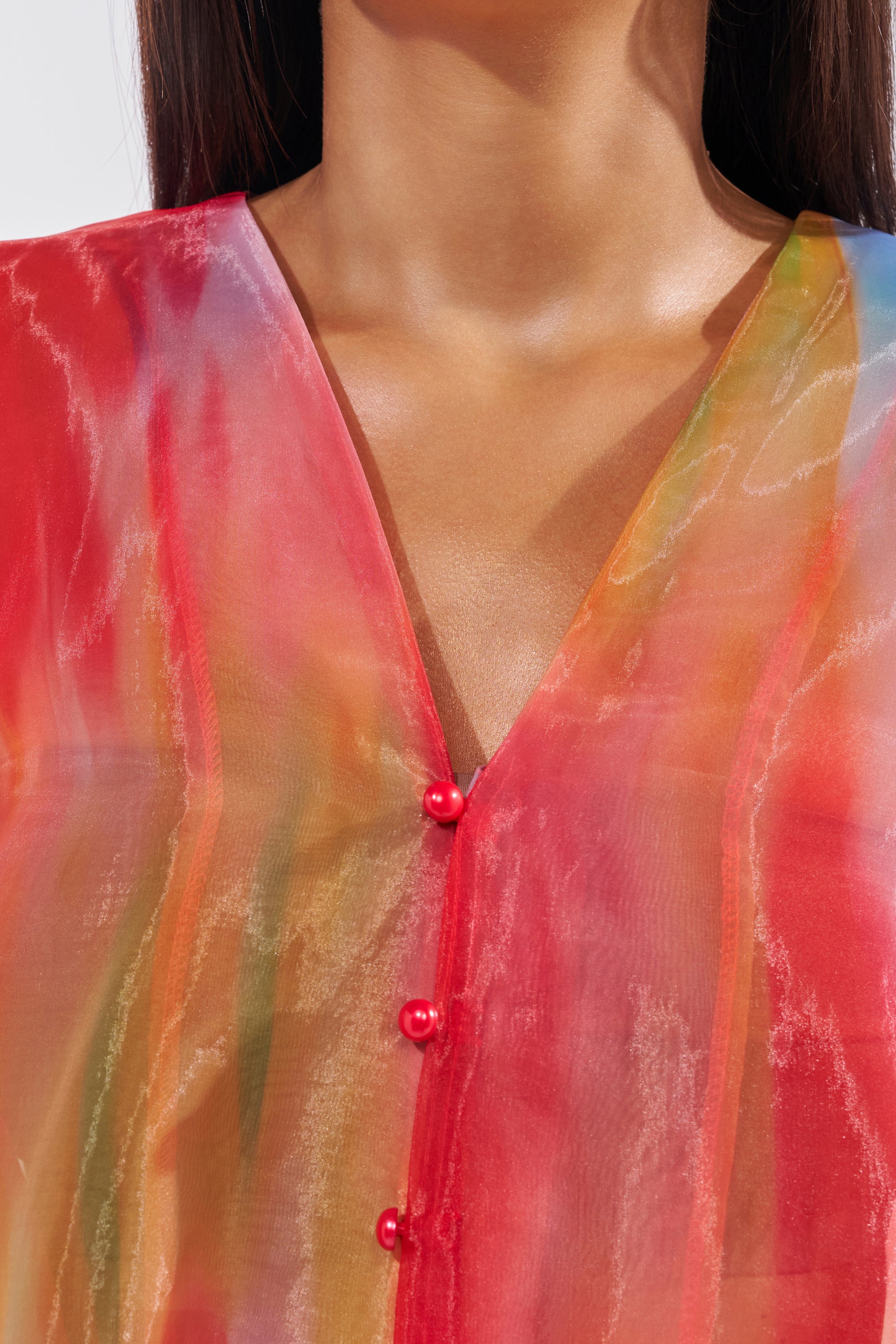 A close-up of a person wearing the INTERESTED IN YOU DUSTER, a sheer organza v-neck blouse with red, pink, and yellow streaks and shiny red buttons. The person's long brown hair is partially visible. 6