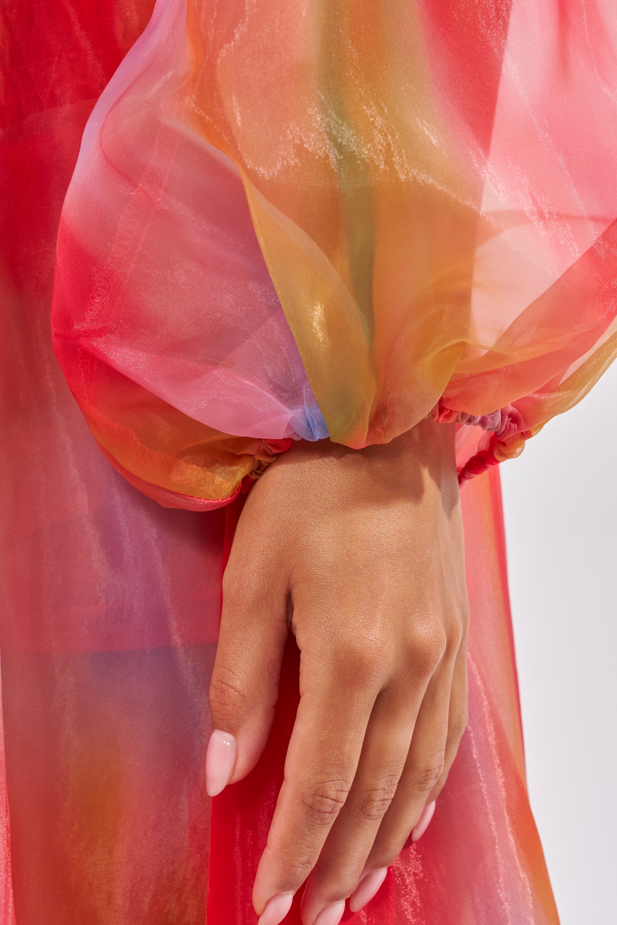 A close-up of a hand with light pink manicured nails rests against the INTERESTED IN YOU DUSTER, a sheer organza piece in vibrant red, orange, pink, and purple. Its shiny texture and longline silhouette create an elegant look. 7