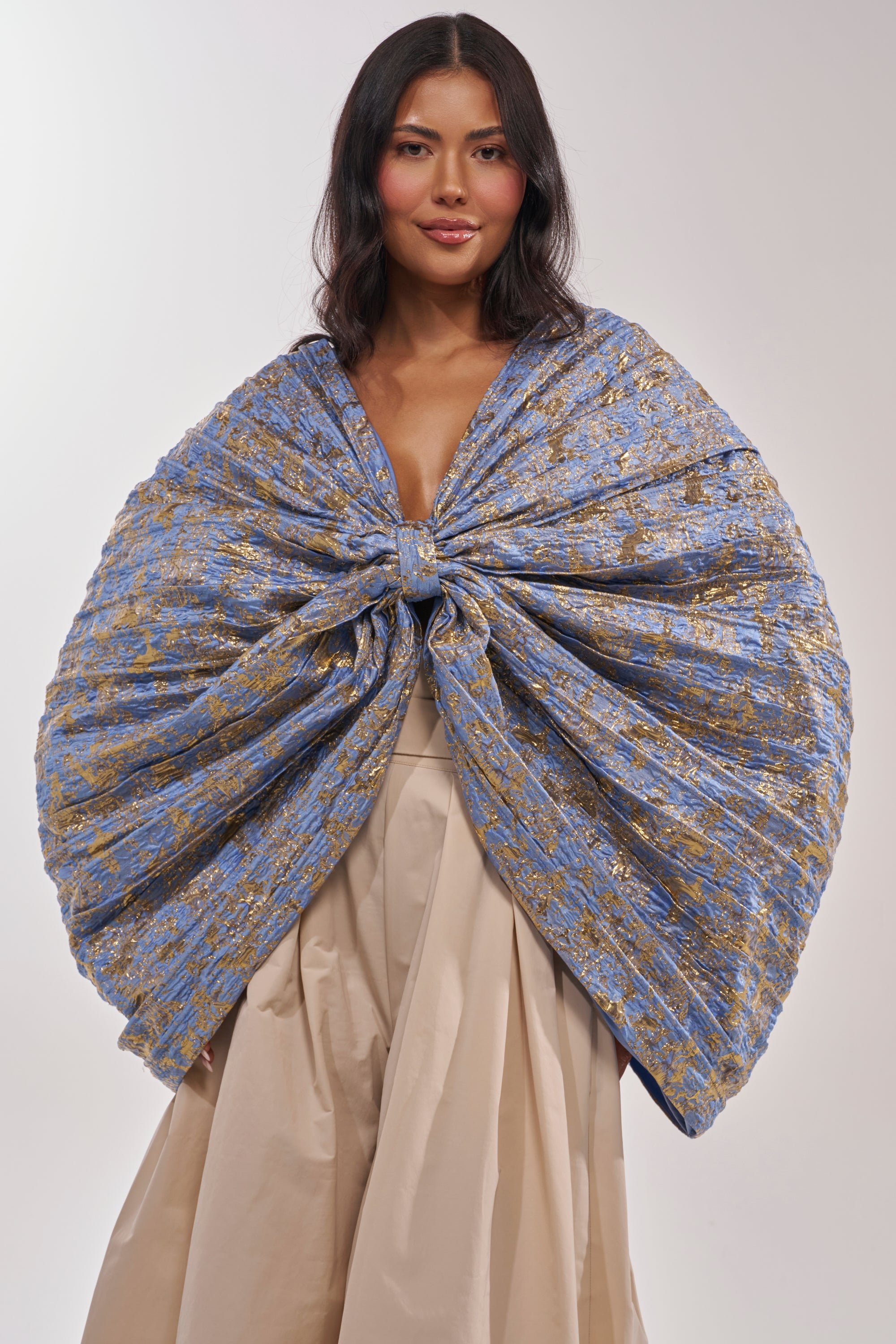 A woman wears the GLORY BROCADE BLOUSE, a blue metallic piece with a statement bow, layered over a beige skirt, standing against a plain light background. 1