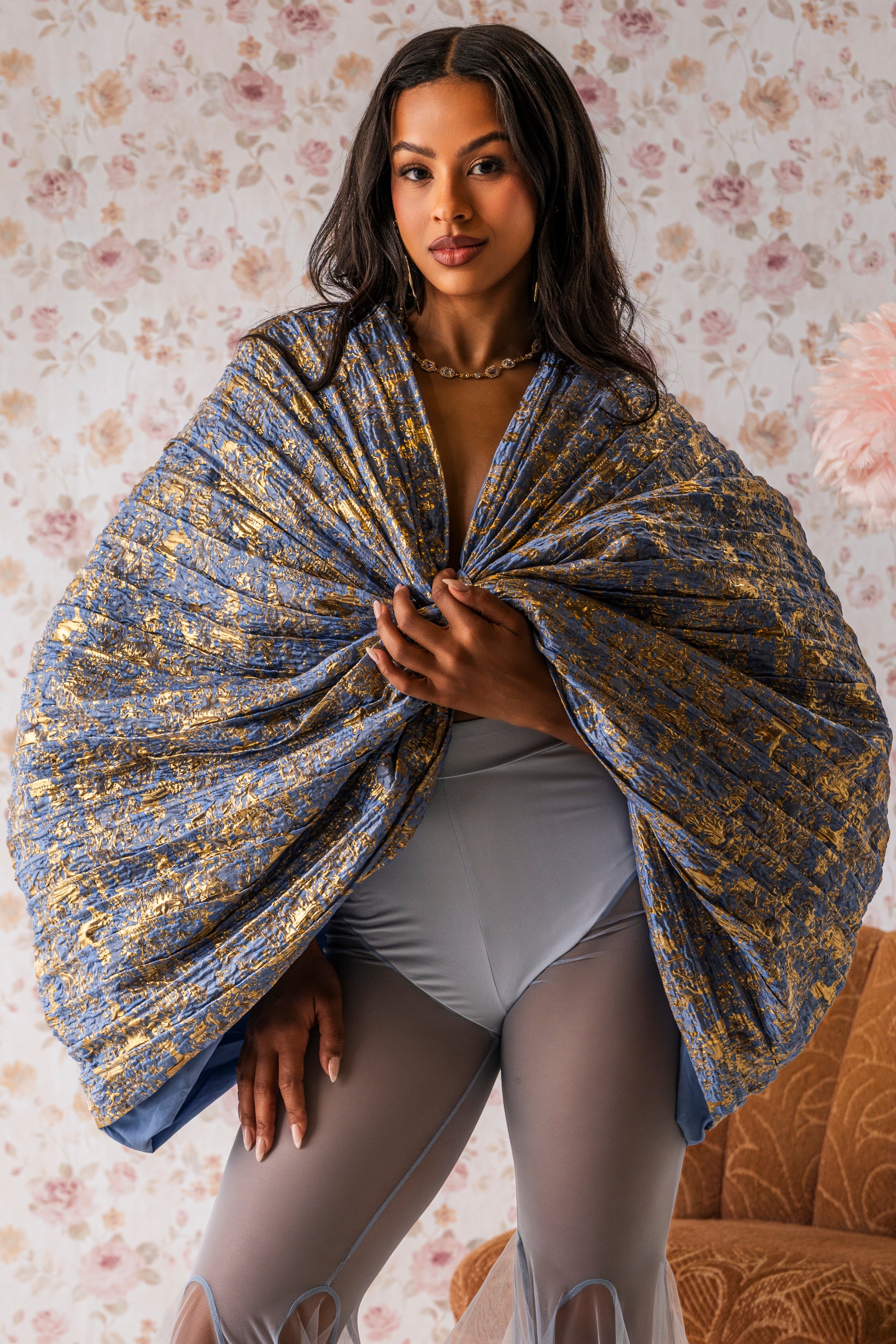 A woman confidently faces the camera in front of floral wallpaper, wearing the dramatic blue metallic GLORY BROCADE BLOUSE draped over her upper body, paired with sheer gray pants. 0