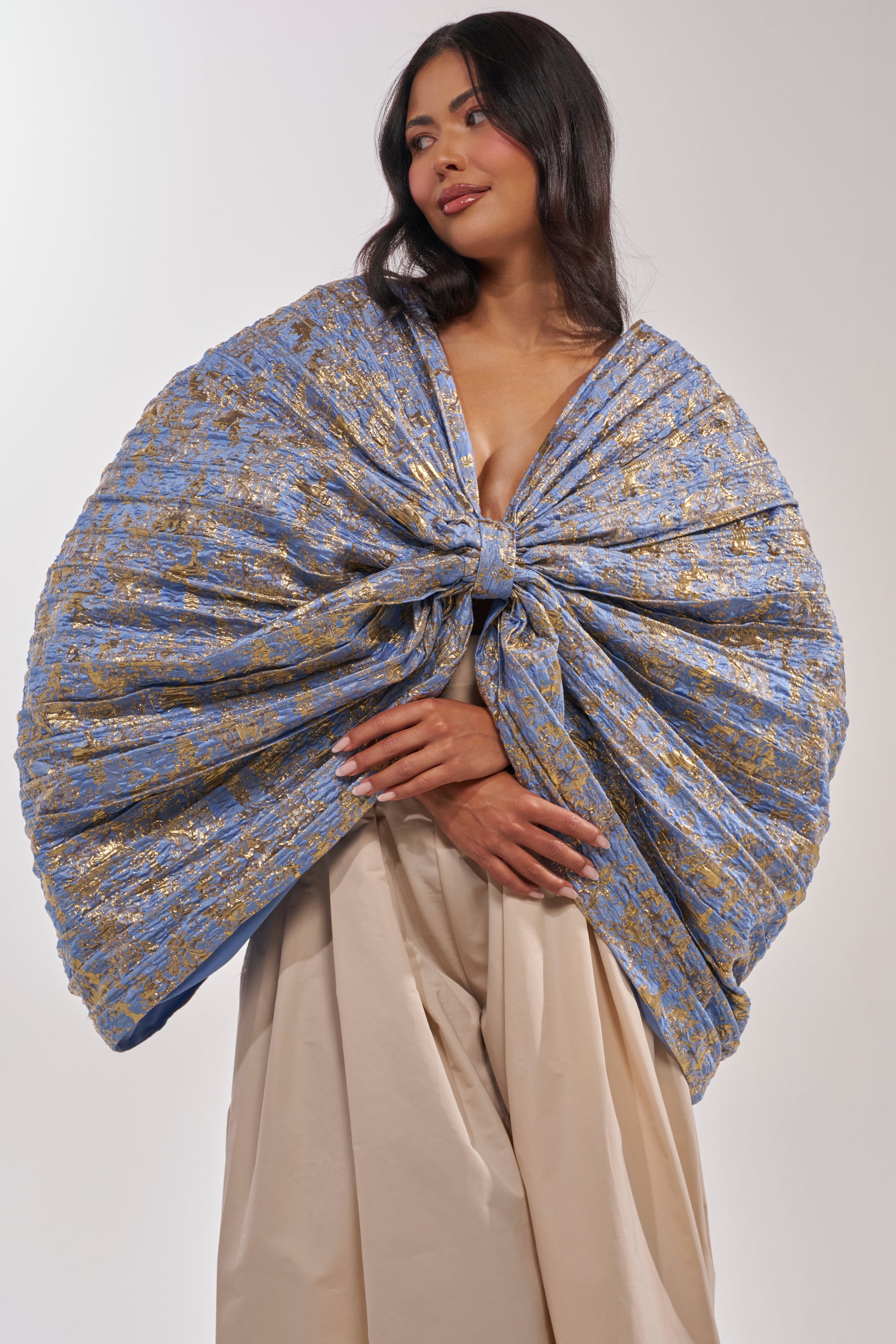 A woman wears the GLORY BROCADE BLOUSE in blue metallic with an oversized bow, paired with cream pants, standing against a white background and smiling softly while looking to the side. 2