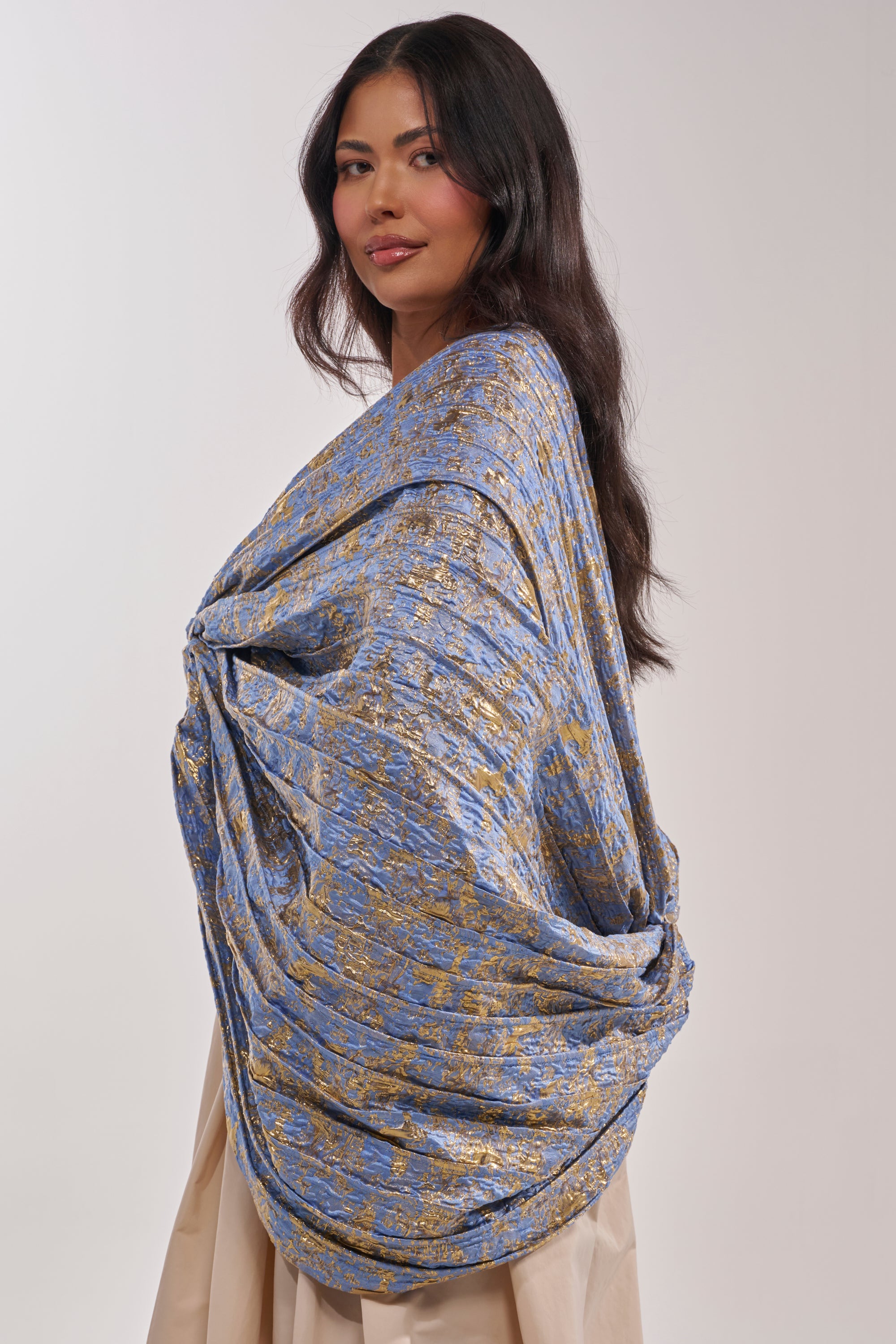 A woman with long dark hair poses in a beige dress styled with the GLORY BROCADE BLOUSE as a voluminous shawl, standing against a plain light background and letting the statement blouse stand out. 3