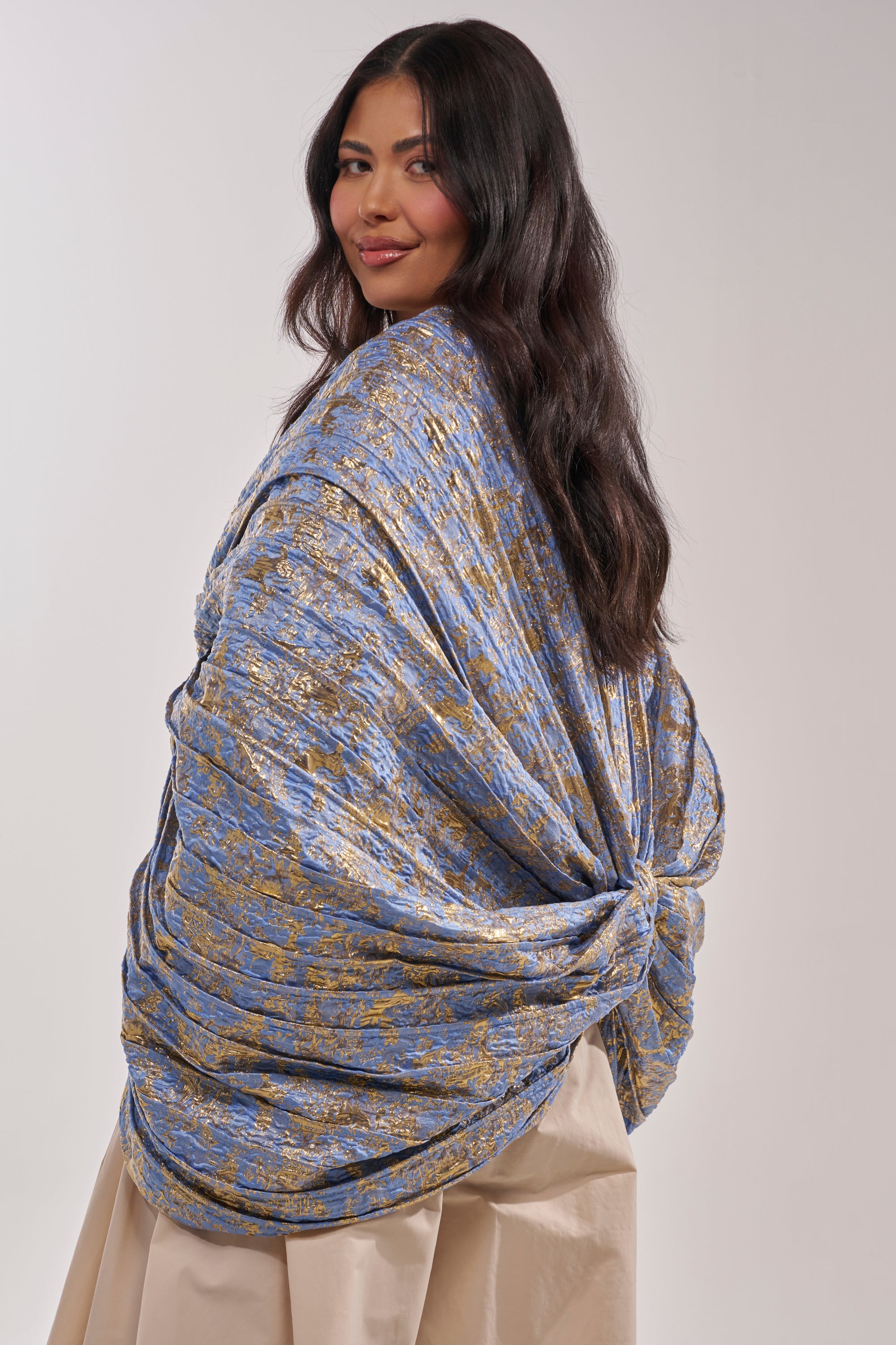 A woman with long, dark hair smiles in the GLORY BROCADE BLOUSE, a loose-fitting blue metallic top with a large bow silhouette at the back, standing against a plain light background. 4