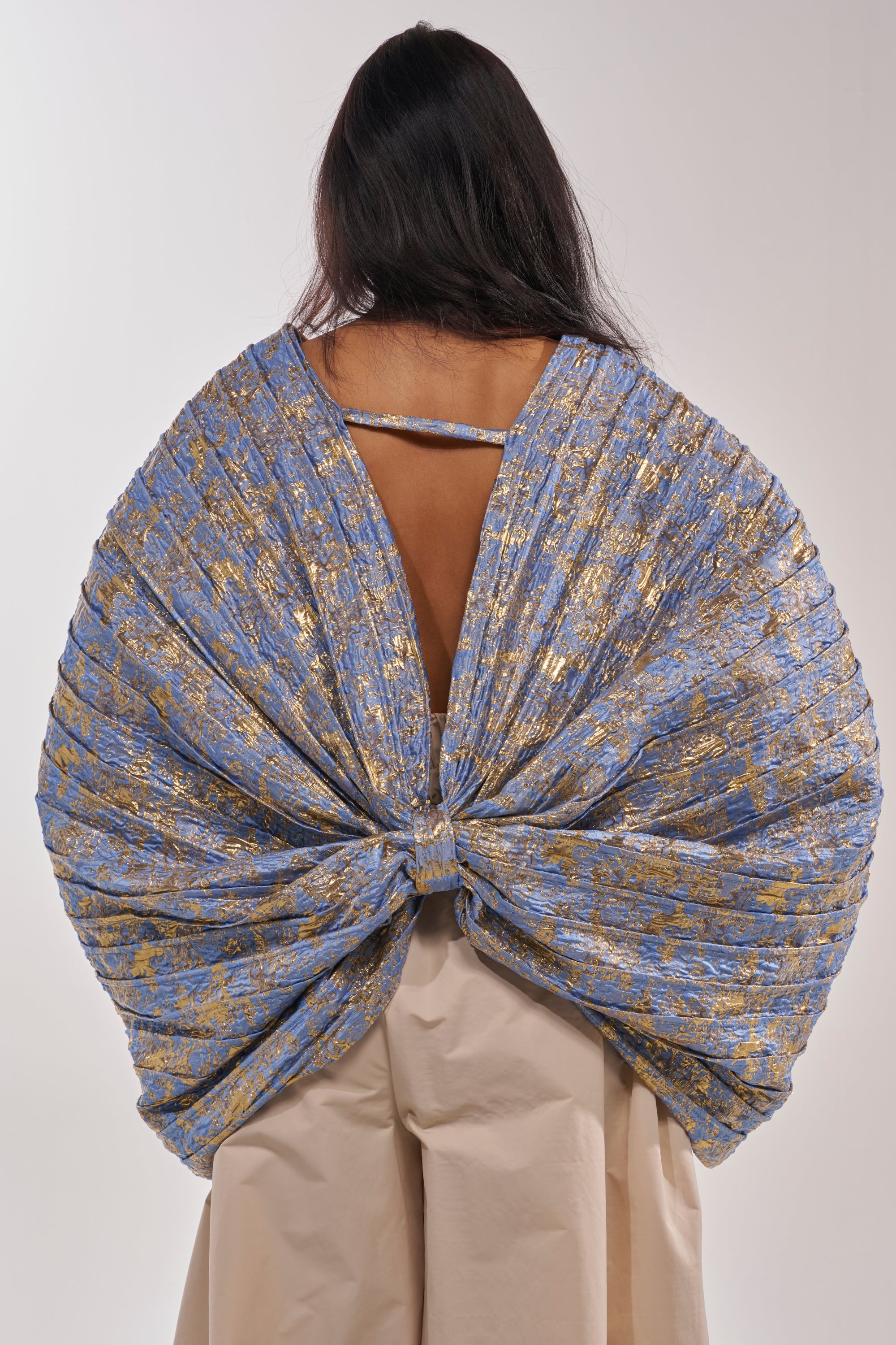 A person stands with their back to the camera, wearing the GLORY BROCADE BLOUSE in dramatic blue metallic fabric with a wide bow silhouette. Their dark hair is loose, and beige pants are visible below the statement piece. 5