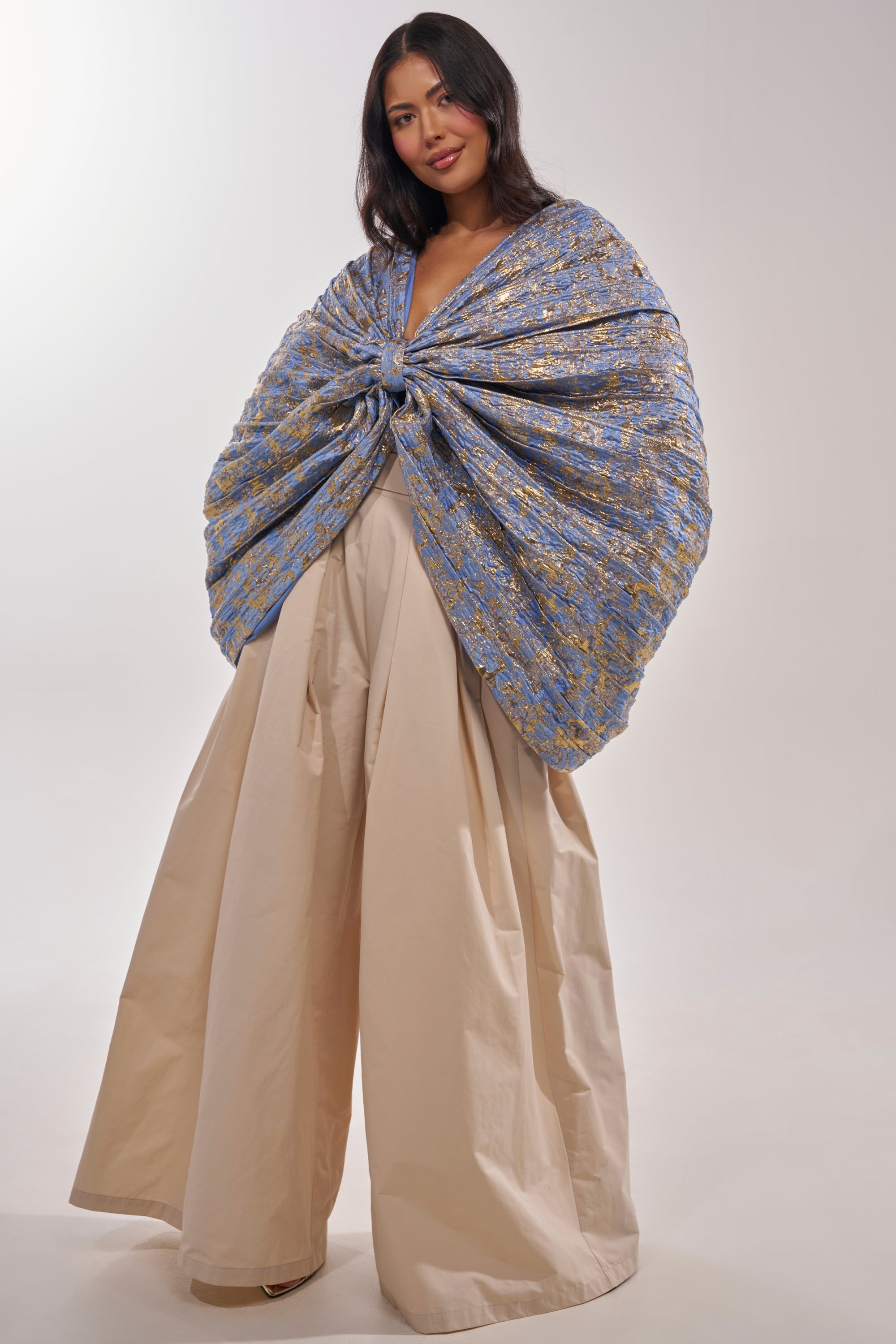 A woman with long dark hair smiles in a studio, wearing the GLORY BROCADE BLOUSE—a dramatic blue metallic top with gold accents, draped and knotted at the chest—paired with wide beige palazzo pants. 6