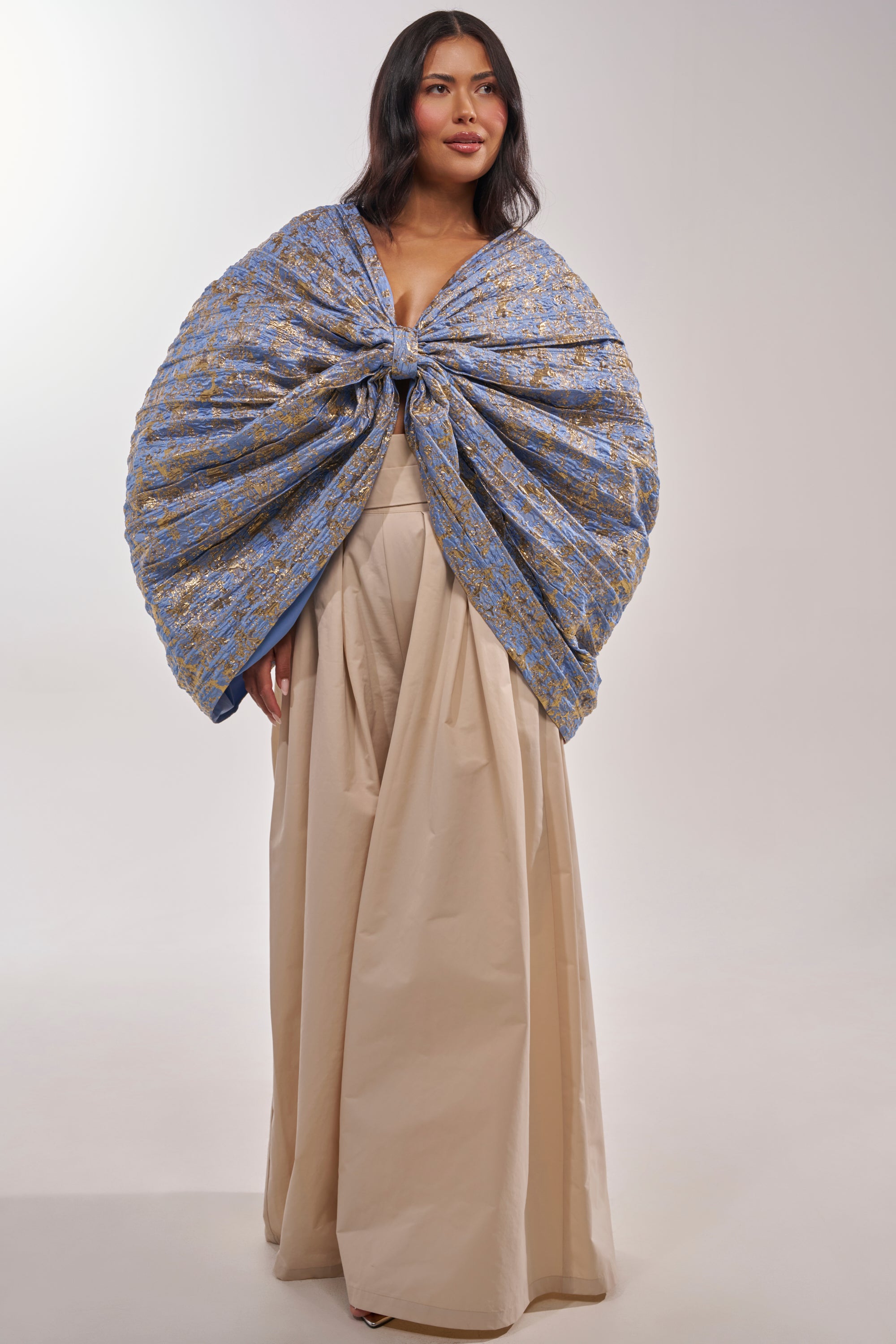 A woman wears the GLORY BROCADE BLOUSE, a blue metallic statement piece with an oversized front bow, paired with a voluminous beige skirt; her long dark hair flows against a plain light background. 7