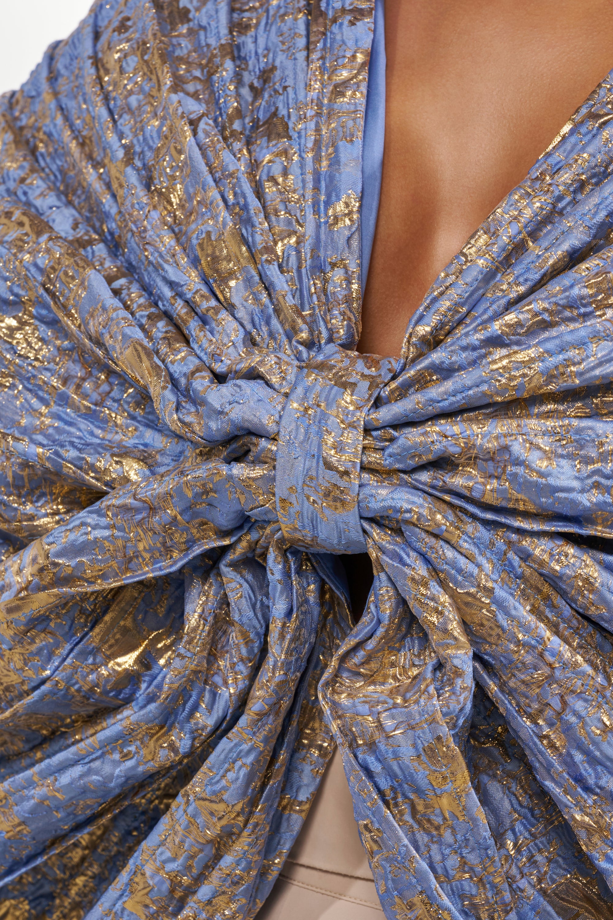 A close-up of a person wearing the GLORY BROCADE BLOUSE in metallic blue and gold, featuring an oversized bow silhouette at the chest and a deep V neckline. 9