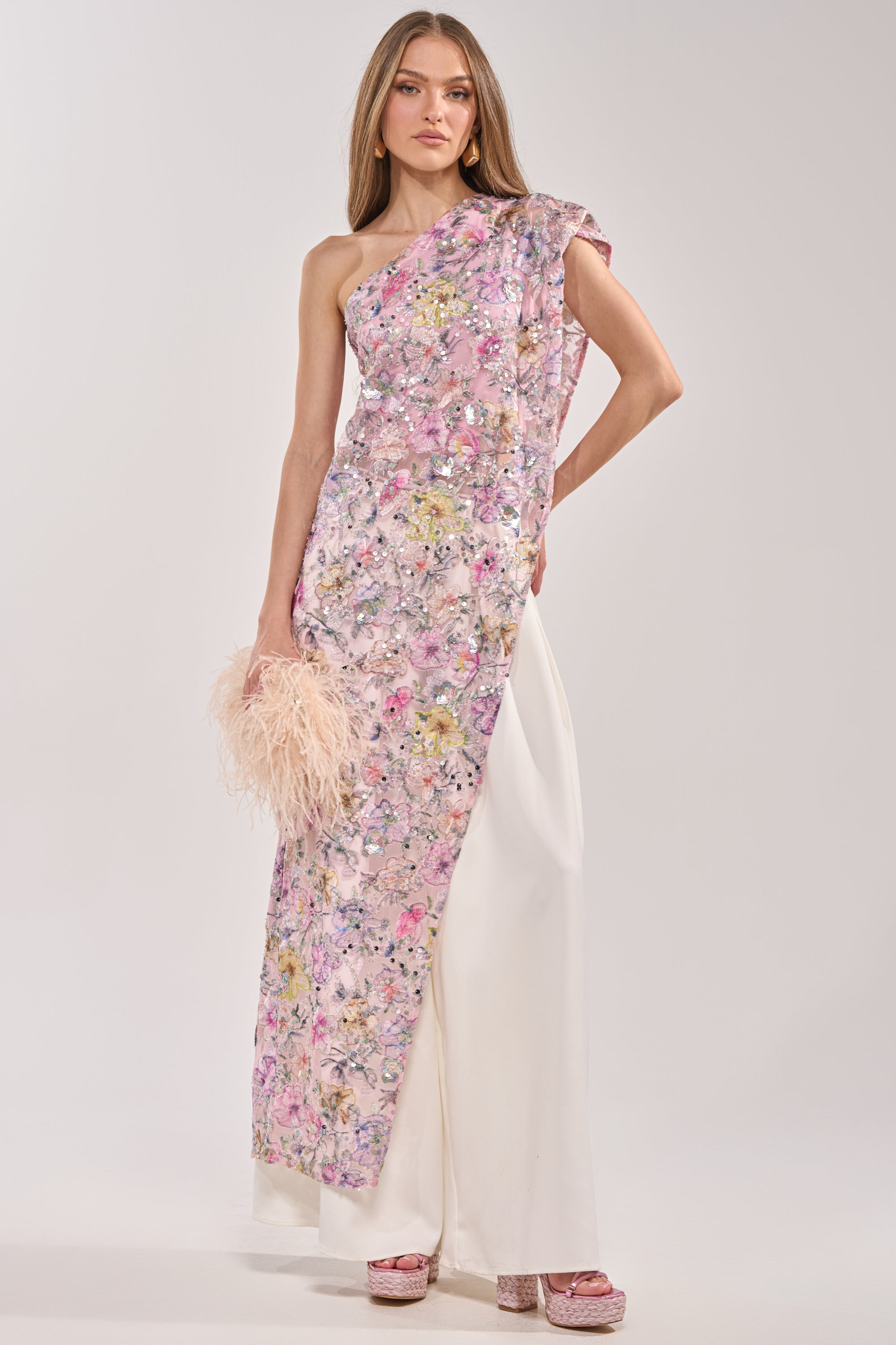 Video of woman modeling the TRINA Floral Elongated Single Shoulder Top in pink and white with wide white pants, holding a pale feathered clutch and platform shoes. Her straight, loose hair frames her face as she poses against a light background.