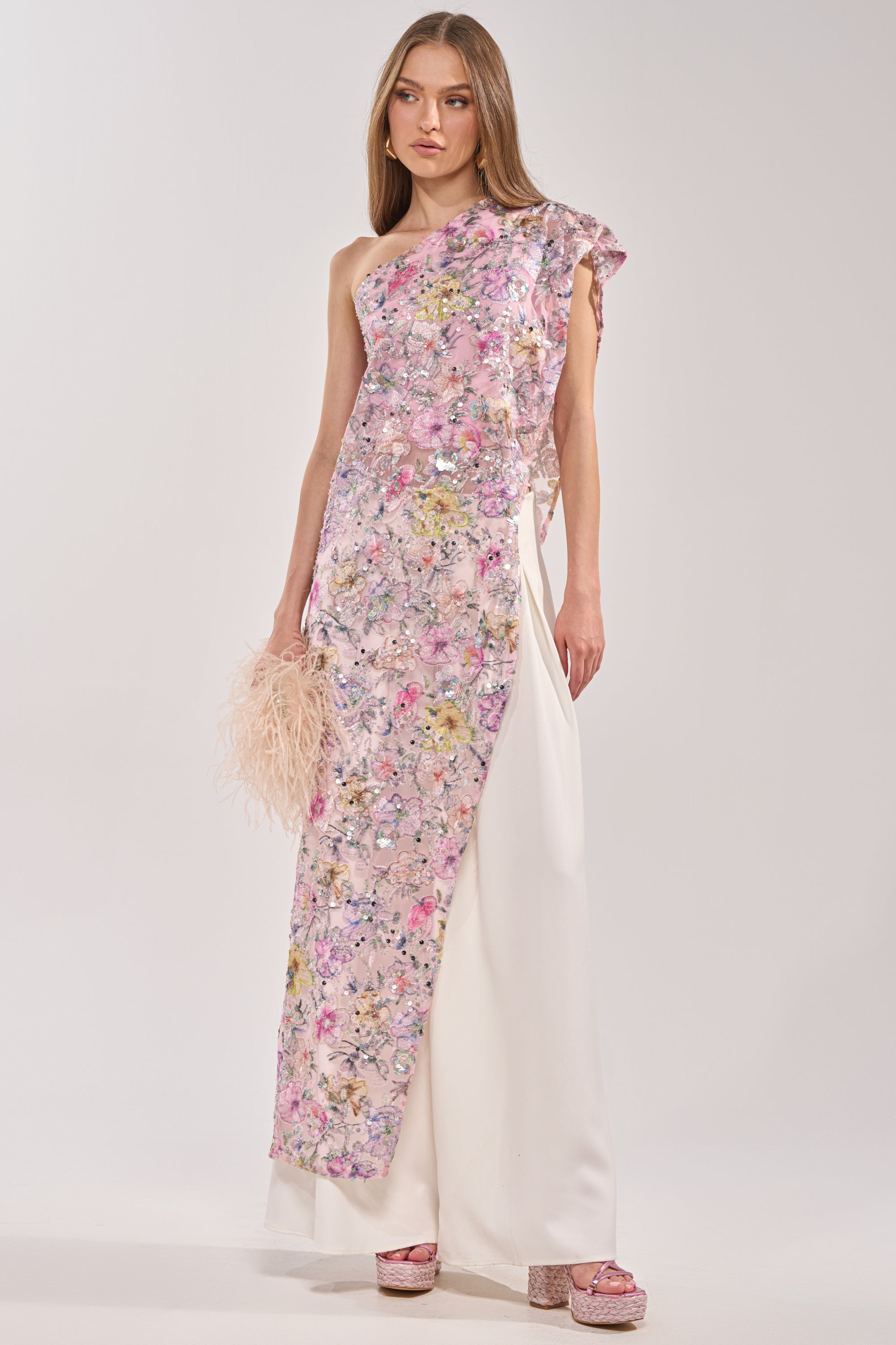 A woman wears the TRINA FLORAL ELONGATED SINGLE SHOULDER TOP with pink mesh floral embroidery and sequins, styled with a floor-length white skirt, pale feathered clutch, and pink platform sandals.