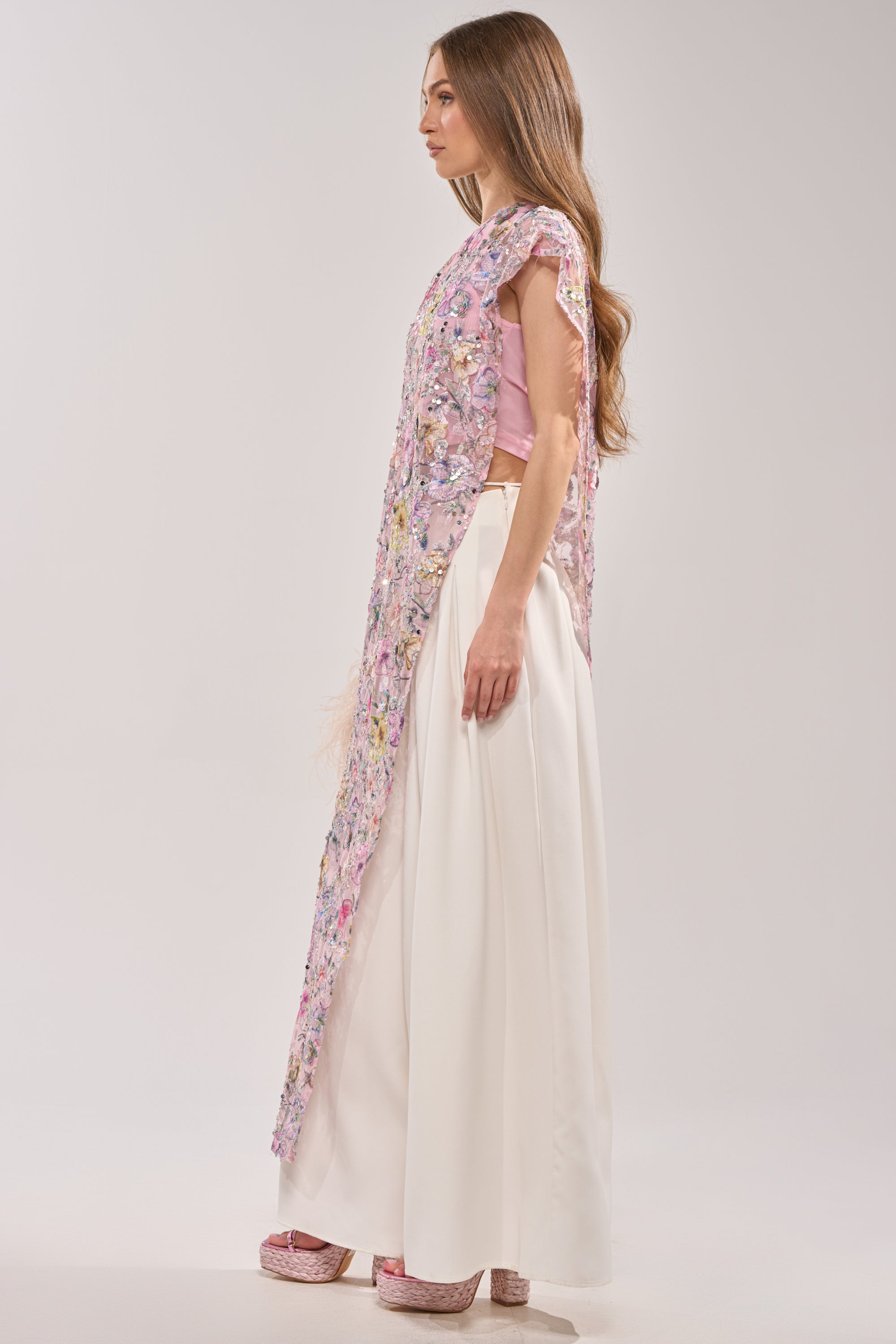 A woman with long brown hair stands sideways against a neutral background wearing the TRINA FLORAL ELONGATED SINGLE SHOULDER TOP, featuring pink mesh floral embroidery and an asymmetrical hem, paired with wide-leg white pants and pink platform shoes.