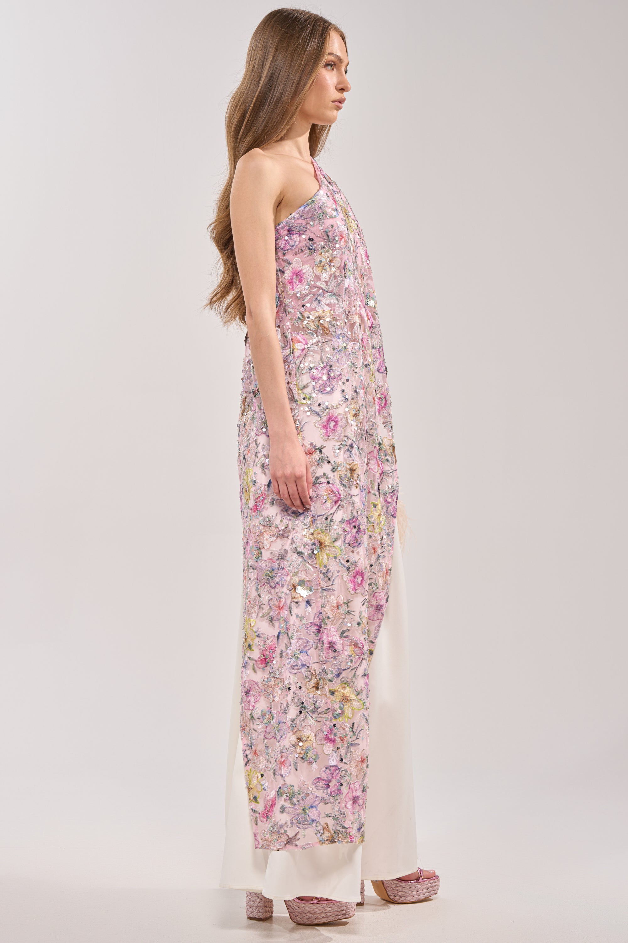 A woman with long brown hair stands in profile against a plain light background, wearing the TRINA FLORAL ELONGATED SINGLE SHOULDER TOP paired with wide-leg white pants and pink platform shoes.