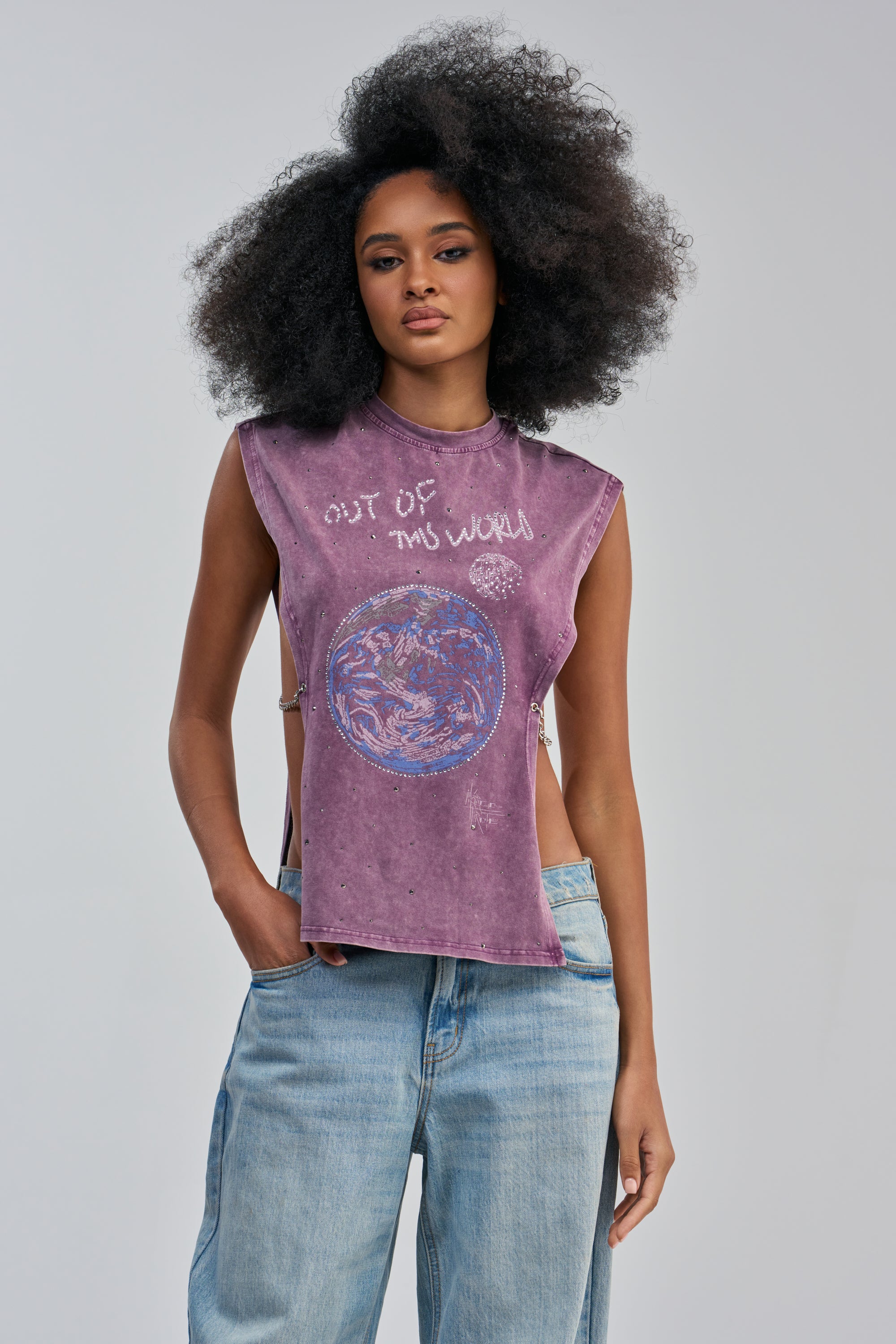 A woman with voluminous curly hair models the WORLDWIDE RHINESTONE EMBELLISHED GRAPHIC TANK—a sleeveless purple streetwear top adorned with “Out of This World” text and a blue-purple planet graphic—styled with light blue jeans against a plain gray background.