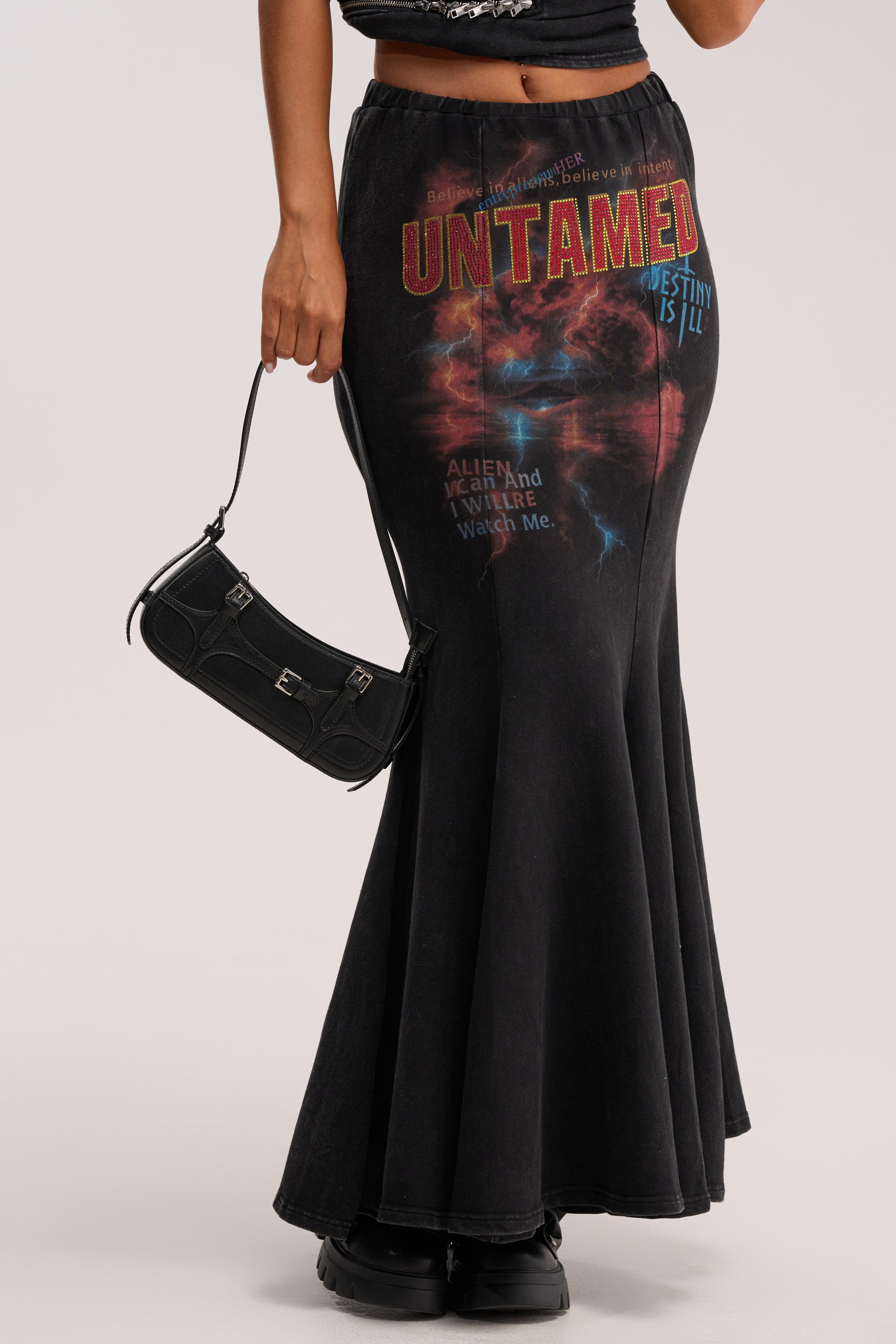 A person styles the UNTAMED FISHTAIL SKIRT, featuring "UNTAMED" text and abstract prints, with a small black handbag and black shoes for an edgy-chic, grunge-inspired look.
