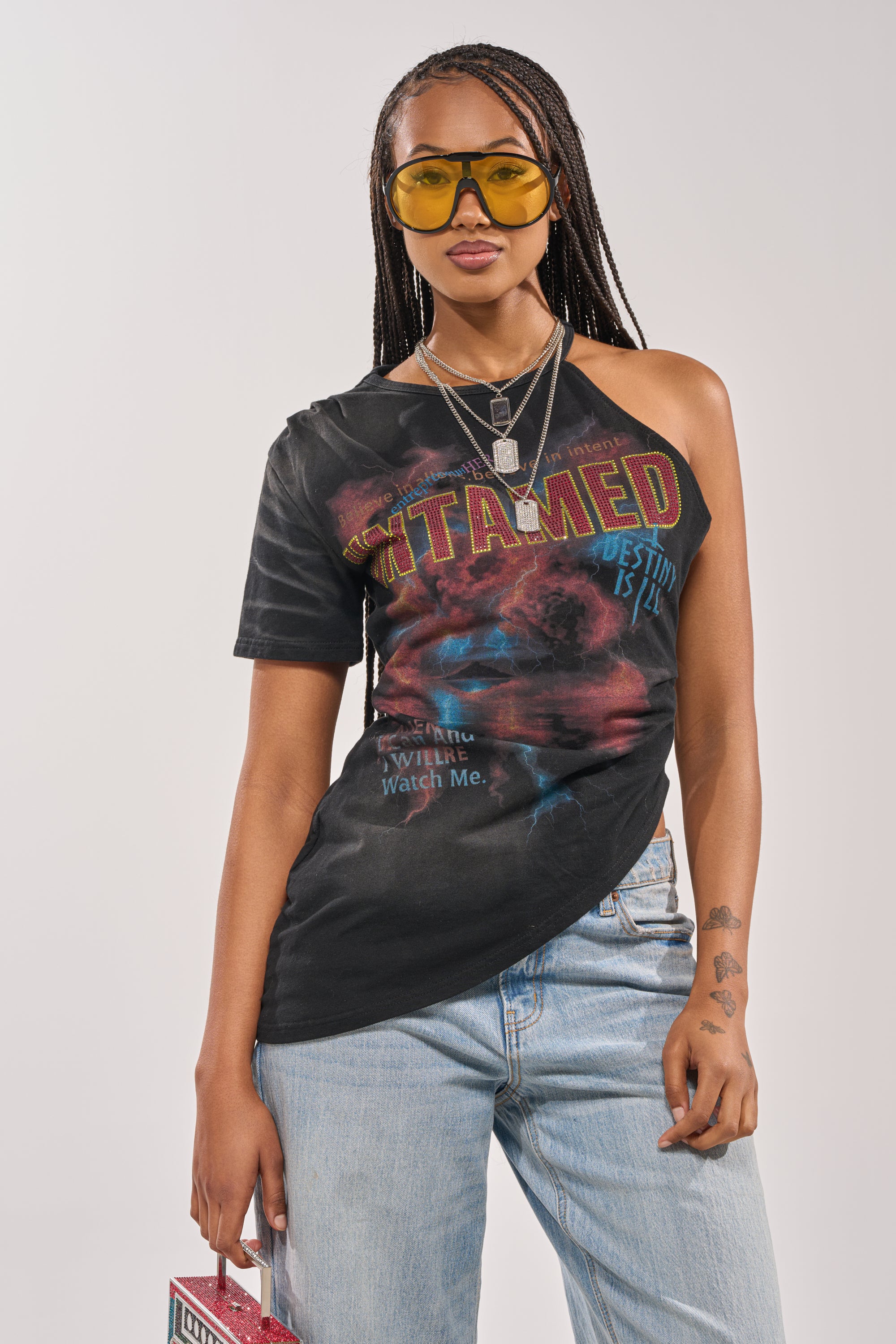 A woman with long braids and yellow-tinted sunglasses wears the UNTAMED SINGLE SLEEVE ASYMMETRICAL TEE in black and light blue jeans, holding a small box that reveals the tattoo on her forearm. 1