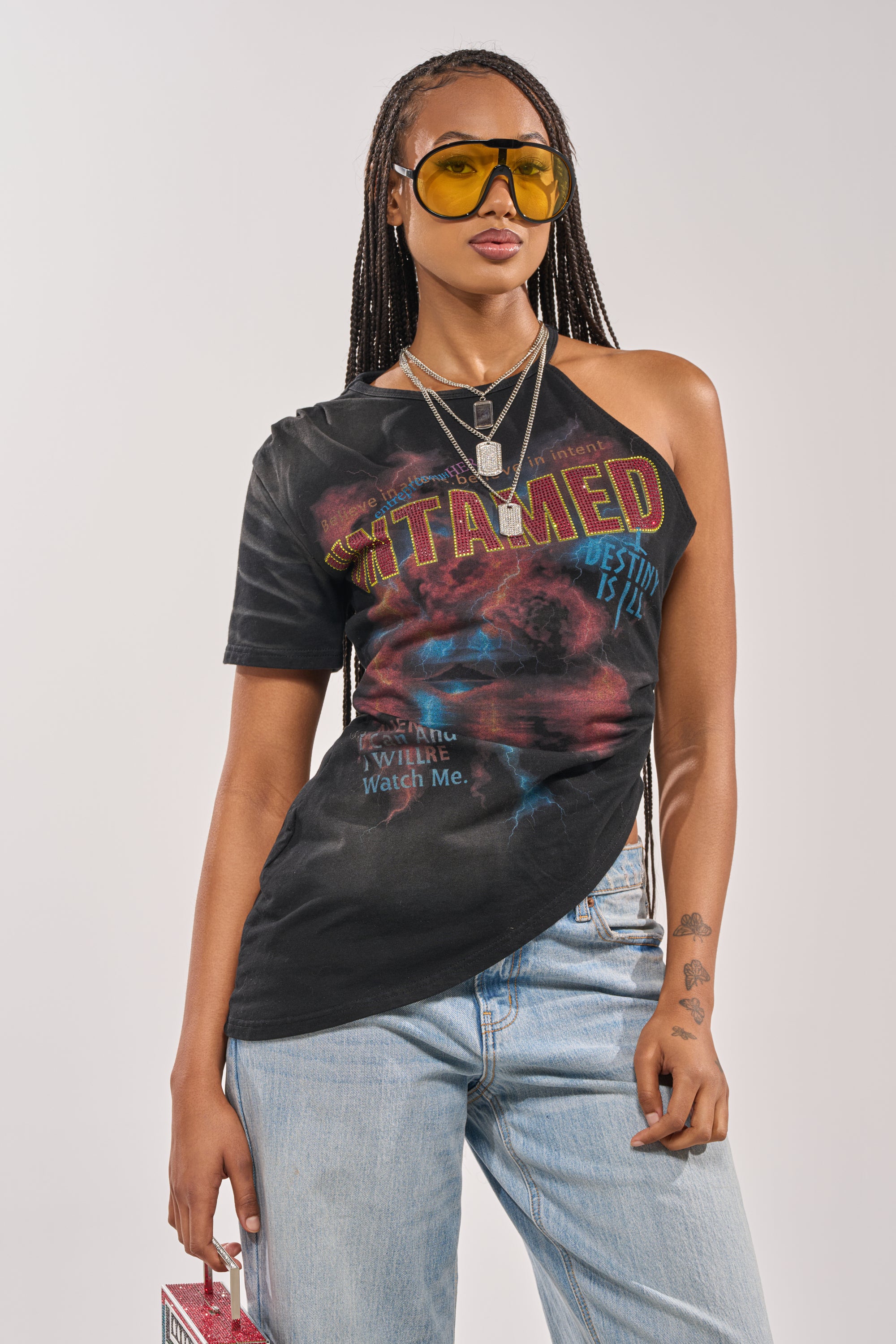 A woman in the UNTAMED SINGLE SLEEVE ASYMMETRICAL TEE, yellow sunglasses, light blue jeans, and layered necklaces stands confidently against a plain background, holding a small pink item in her left hand. 2