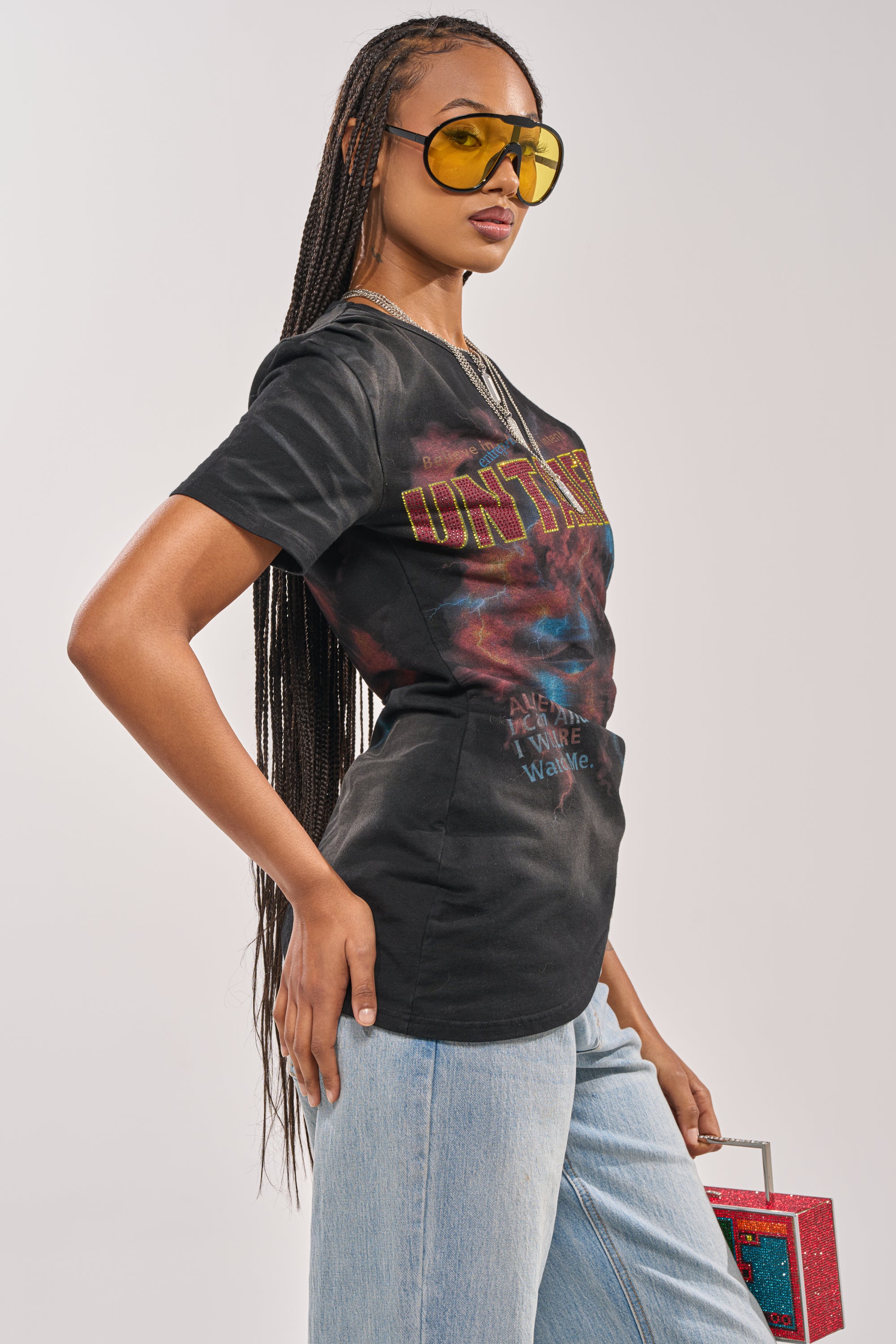 A woman with long braided hair wears yellow-tinted sunglasses, light blue jeans, and the UNTAMED SINGLE SLEEVE ASYMMETRICAL TEE. She stands in a side pose, holding a small red glittery handbag against a plain background. 3