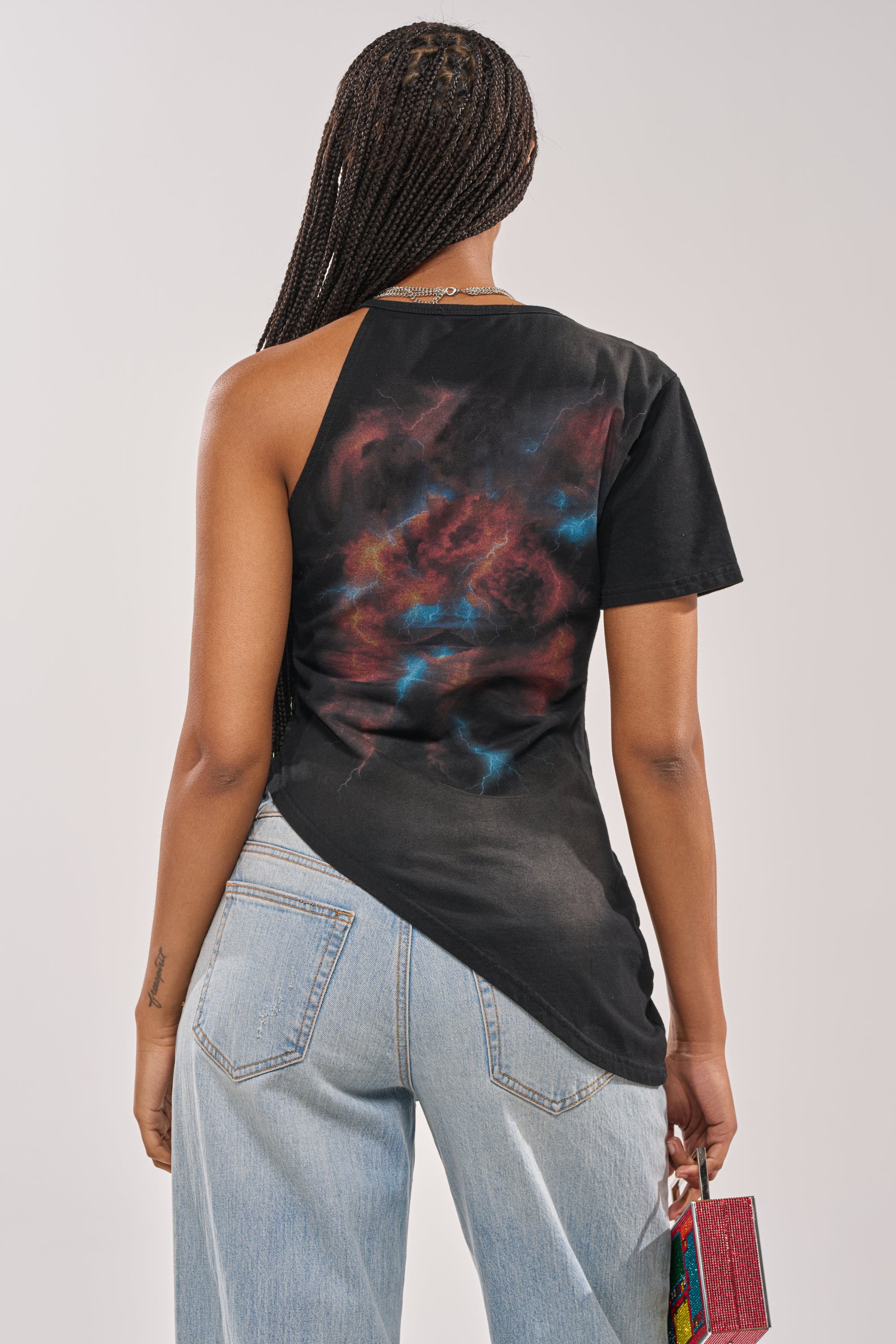 A person with long braids, seen from behind, wears the UNTAMED SINGLE SLEEVE ASYMMETRICAL TEE in black with a colorful galaxy print on the back, paired with light blue jeans and holding a small purse in their left hand. 5