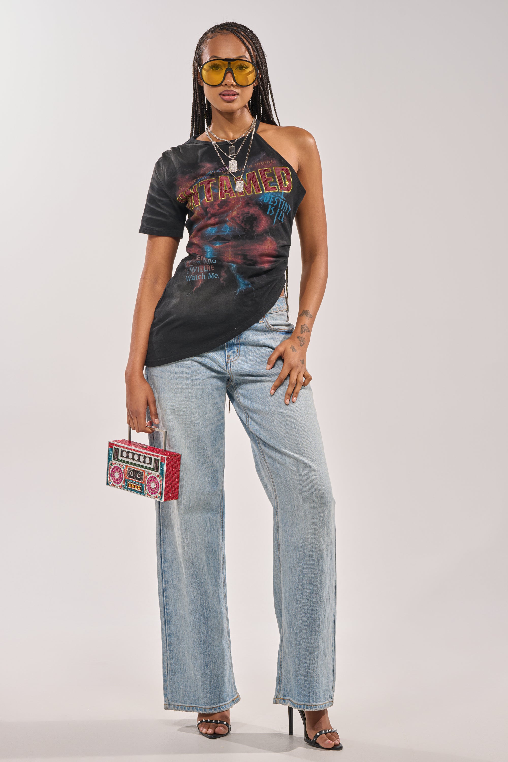 A woman with braids wears yellow sunglasses, the UNTAMED SINGLE SLEEVE ASYMMETRICAL TEE, light blue wide-leg jeans, black heels, holds a red boombox-shaped purse, and finishes her outfit with layered necklaces. 6