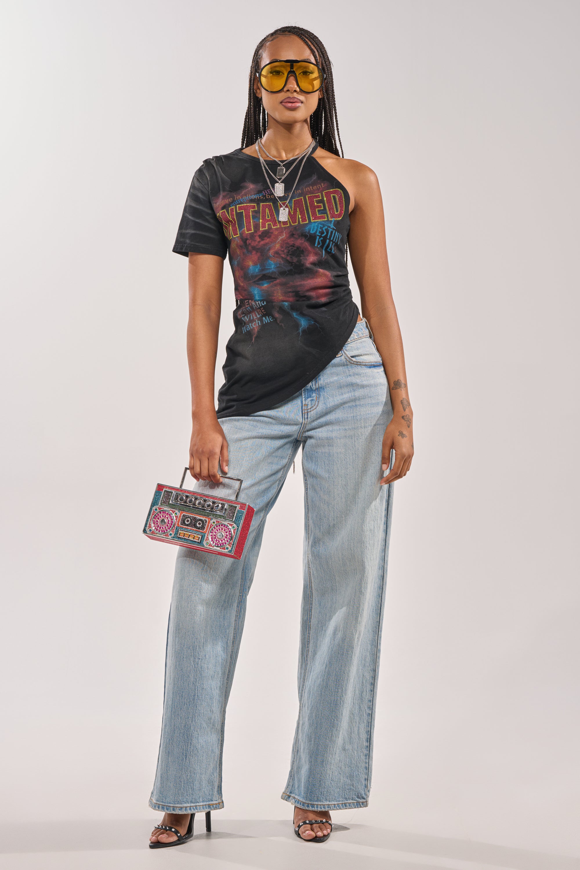 A person with long braids wears yellow sunglasses, the UNTAMED SINGLE SLEEVE ASYMMETRICAL TEE, light blue wide-leg jeans, black heels, and holds a colorful rectangular clutch. 8