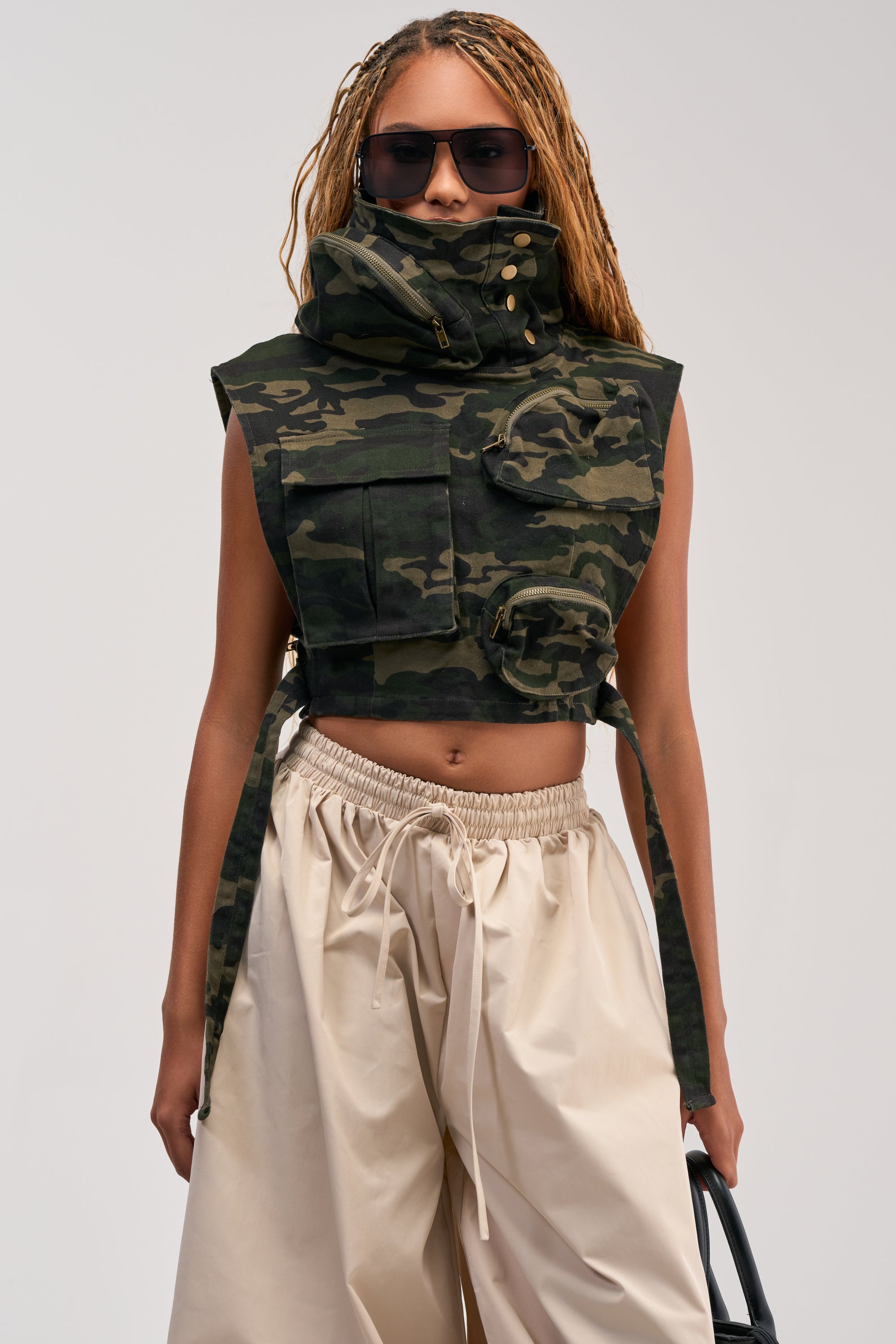 A person wearing sunglasses, the KADEN CARGO SLEEVELESS TOP IN CAMO styled as a streetwear utility vest with large pockets, and loose beige pants stands against a plain background, holding a bag.