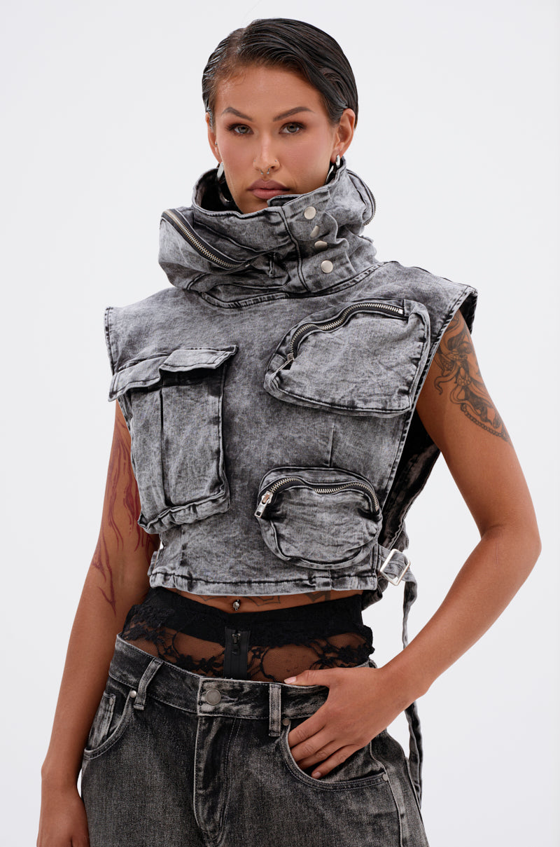 A woman confidently poses against a plain white background wearing the MODEL BEHAVIOR DENIM TOP—a sleeveless, cropped gray denim vest with multiple large pockets and zippers—paired with matching acid-wash black denim jeans and a black lace top.